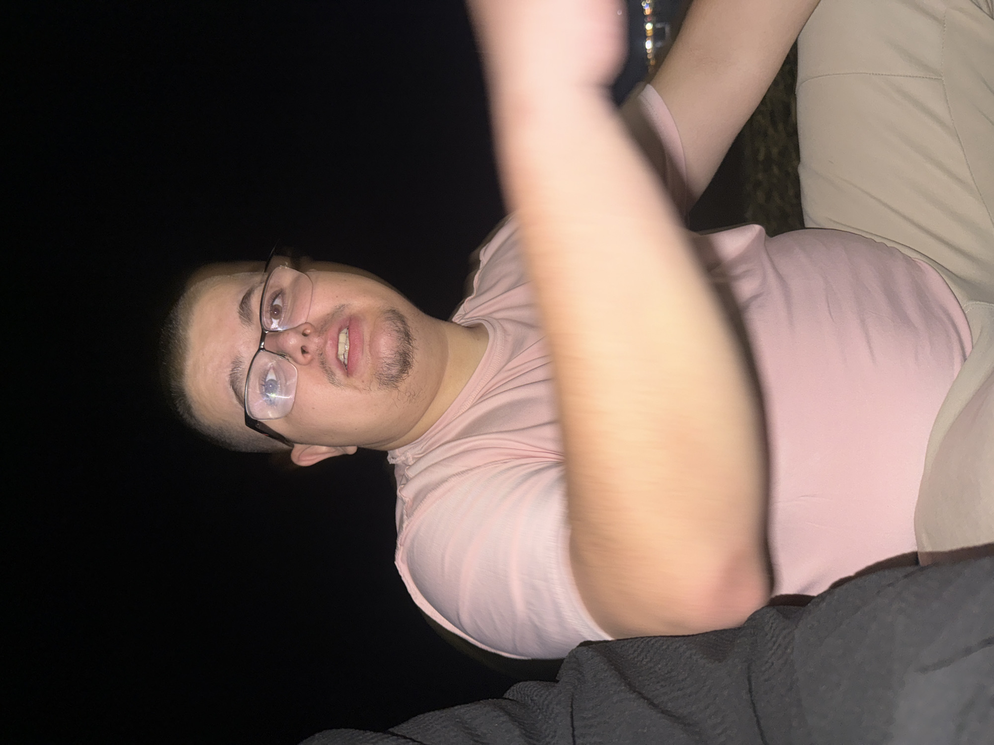 This image depicts a nighttime outdoor scene in the city of Varna, Bulgaria. The central figure is a male adult with a light to medium complexion, short dark hair, and a goatee, wearing clear-framed glasses. He is dressed in a light pink short-sleeved t-shirt and light beige trousers. His mouth is slightly agape, revealing teeth or dental braces, and he is looking directly at the camera. His left arm is extended towards the foreground, appearing significantly blurred due to motion or close proximity to the lens. To the immediate left of the central figure, a second individual is partially visible, wearing a dark, textured garment that appears to be a sweater or jacket. The background is predominantly dark, with indistinct, blurred lights, consistent with distant city lights or lights across a body of water, visible in the lower right corner of the frame. The subjects are illuminated by a strong, direct light source, likely a camera flash, indicating a close-range photograph taken in low-light conditions.