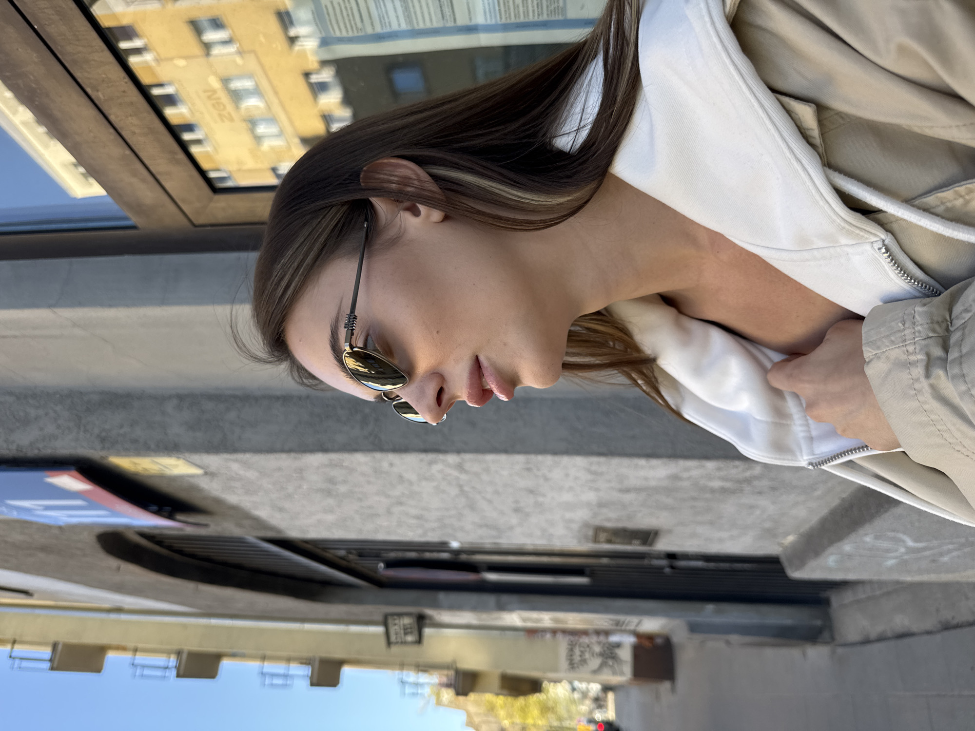 A young woman is pictured in a medium shot, seemingly from a low angle. She wears sunglasses and a light-colored jacket over a white hoodie. The background reveals a street scene in Warsaw, Poland, with buildings and urban infrastructure visible. She is standing near an urban element, likely a ventilation grating.