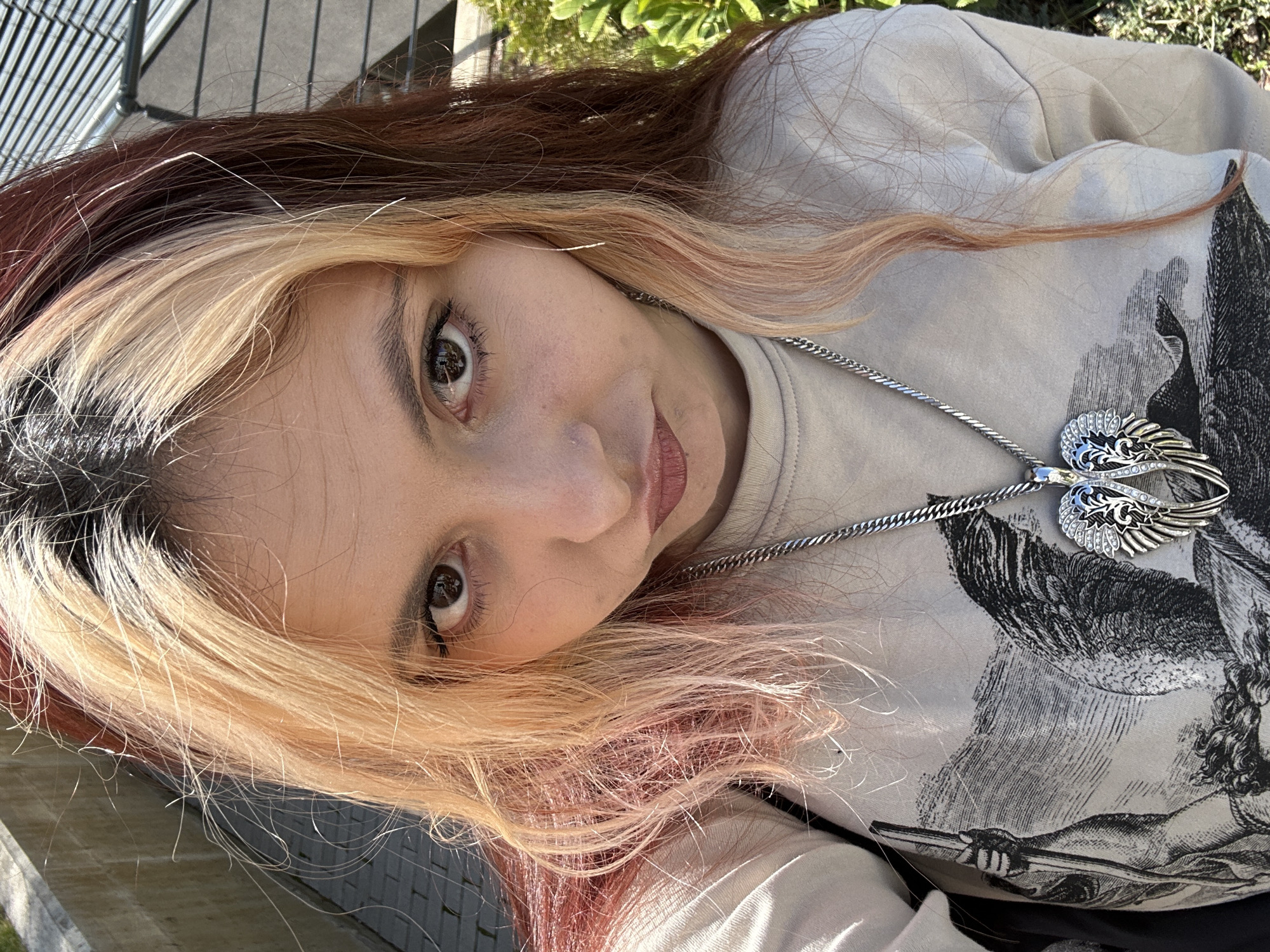 A young woman with light skin and brown eyes is oriented at an angle towards the camera. She has multi-colored hair with brown, blonde, and pink hues. She wears a light-colored top with a graphic design on the front and a silver necklace with a pendant featuring wings. The background includes greenery and an outdoor structure with a metallic grid. The location cannot be confirmed as Sofia, Bulgaria.