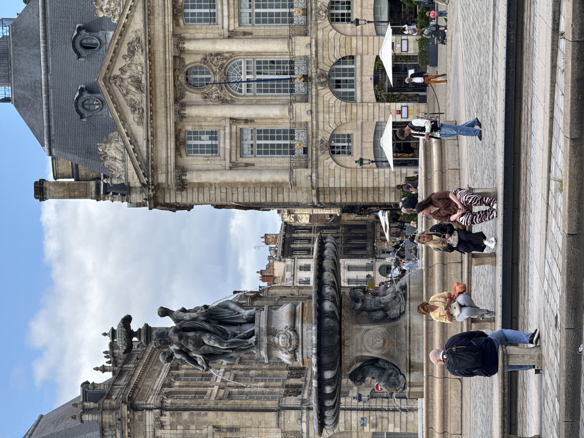 The image depicts a view of the Place de la Bourse in Bordeaux, France. In the foreground, a large ornate fountain with bronze sculptures of figures is visible, with water spraying from it. Several people are seated on stone benches around the fountain, facing away from the camera or looking at their phones. A man in a black backpack and blue jeans sits on a bench on the left, while three women sit on a bench to the right of the fountain. Behind the fountain, a grand stone building with arched windows and classical architectural elements dominates the background. To the right of this building, another similar building is partially visible. Several white umbrellas indicating outdoor seating for a cafe or restaurant are set up in front of the buildings. People are walking in the background. The sky is overcast with scattered clouds.