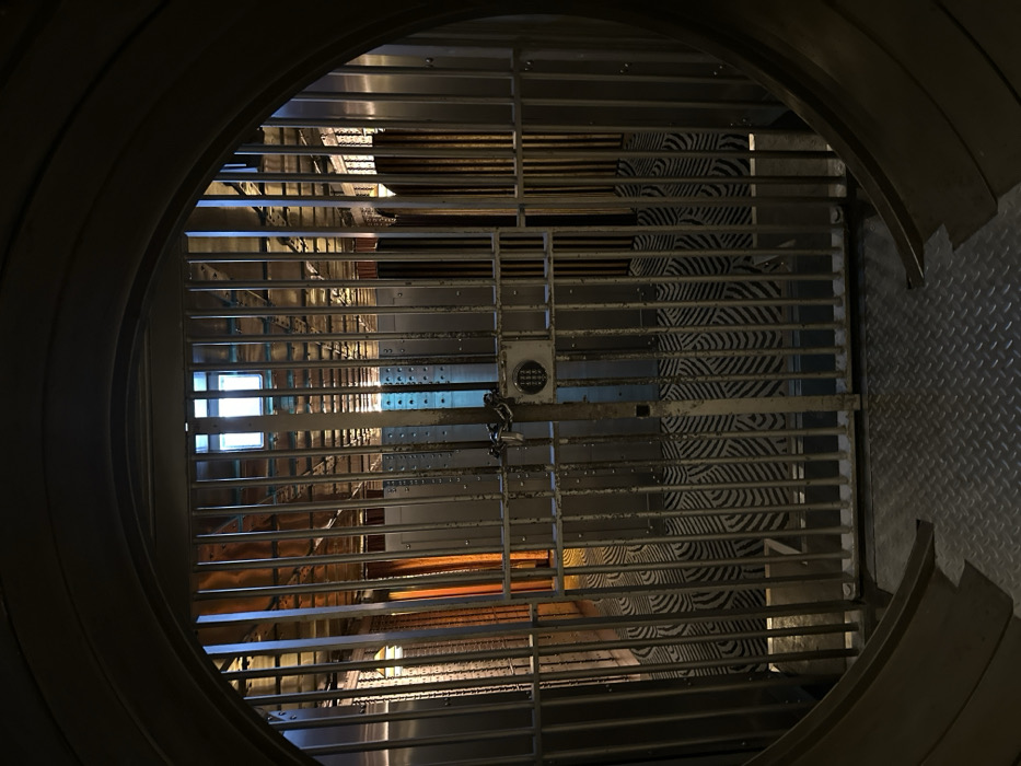 The image depicts the interior view of "The Vault" bar, located within The Ned hotel, formerly the Midland Bank building, in the City of London, United Kingdom.
The foreground consists of a dark grey diamond plate metal floor, which transitions into a large, circular opening of a bank vault. Centered within this vault opening is a robust, silver-colored metal grated gate, composed of numerous vertical bars and a thicker central vertical bar. Attached to this central bar is a black digital keypad lock, secured with a silver chain wrapped around the bar.
Beyond the grated gate, a long, corridor-like interior space is visible. The floor of this space is covered with a distinctive patterned carpet featuring repeating, curved, wave-like or scalloped designs in dark and light grey tones. The walls of the corridor are lined with polished metal panels, some appearing as individual safe deposit boxes or secure storage units, reflecting various light sources.
Illumination within the vault interior comes from multiple sources: a prominent rectangular window-like light fixture at the far end emits a bright white light. Vertical strips of warm orange-yellow light illuminate panels on the left side of the corridor, while cooler blue light is visible in other sections. The ceiling features exposed metal framework and conduits. No individuals or active interactions are present within the scene.