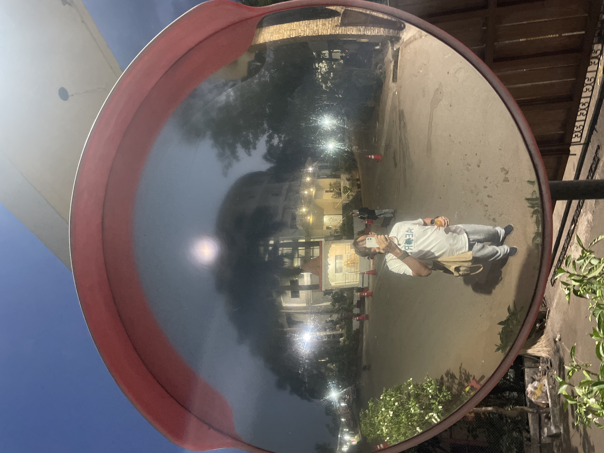 The image shows a convex mirror reflecting a street scene at night. A person is taking a photo of themself in the mirror. The reflection includes a building with multiple lights and a small Hindu temple. Orange traffic cones are placed on the roadside. The surroundings are residential with greenery and typical urban infrastructure visible. Given the temple and general architecture, it's likely located in Mumbai, India.
