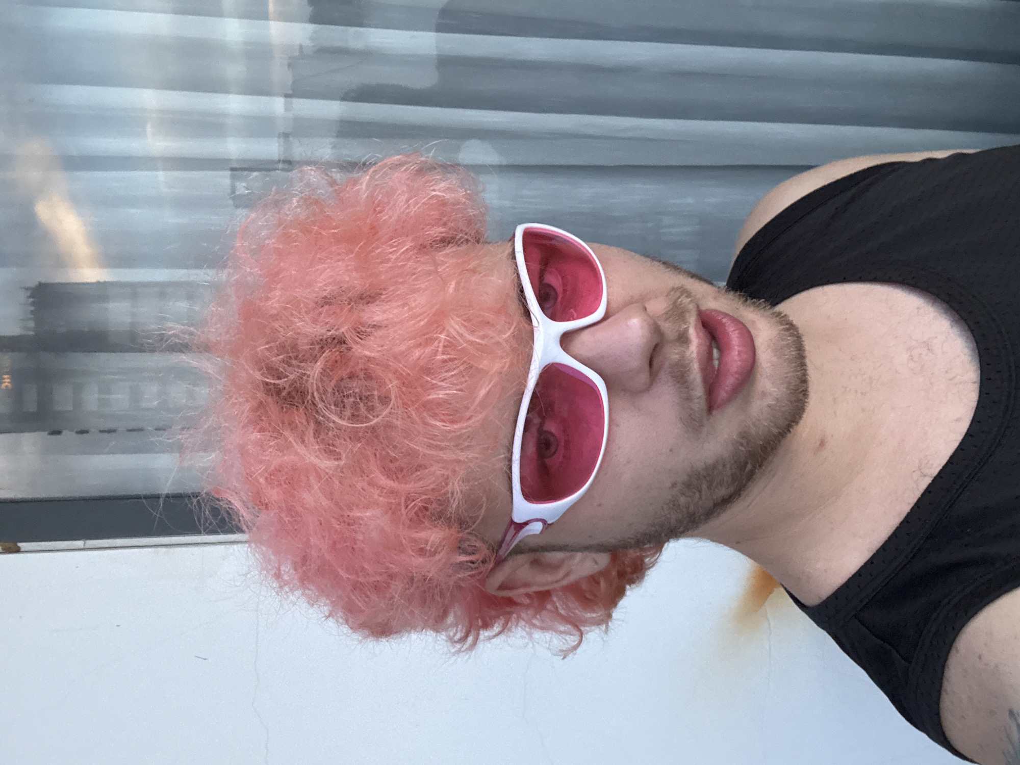 A close-up, eye-level, medium shot features a person with bright pink, curly hair. They are wearing a black tank top and bright pink sunglasses with white frames. The person's mouth is slightly open, revealing their teeth. The background is divided, with a textured white wall on the left and a light blue curtain on the right, behind which a blurred city skyline with high-rise buildings is visible. The lighting suggests it is daytime, possibly late afternoon due to the warm light reflecting on the curtain. There is no visible indication of the specific location within Da Nang, Vietnam.