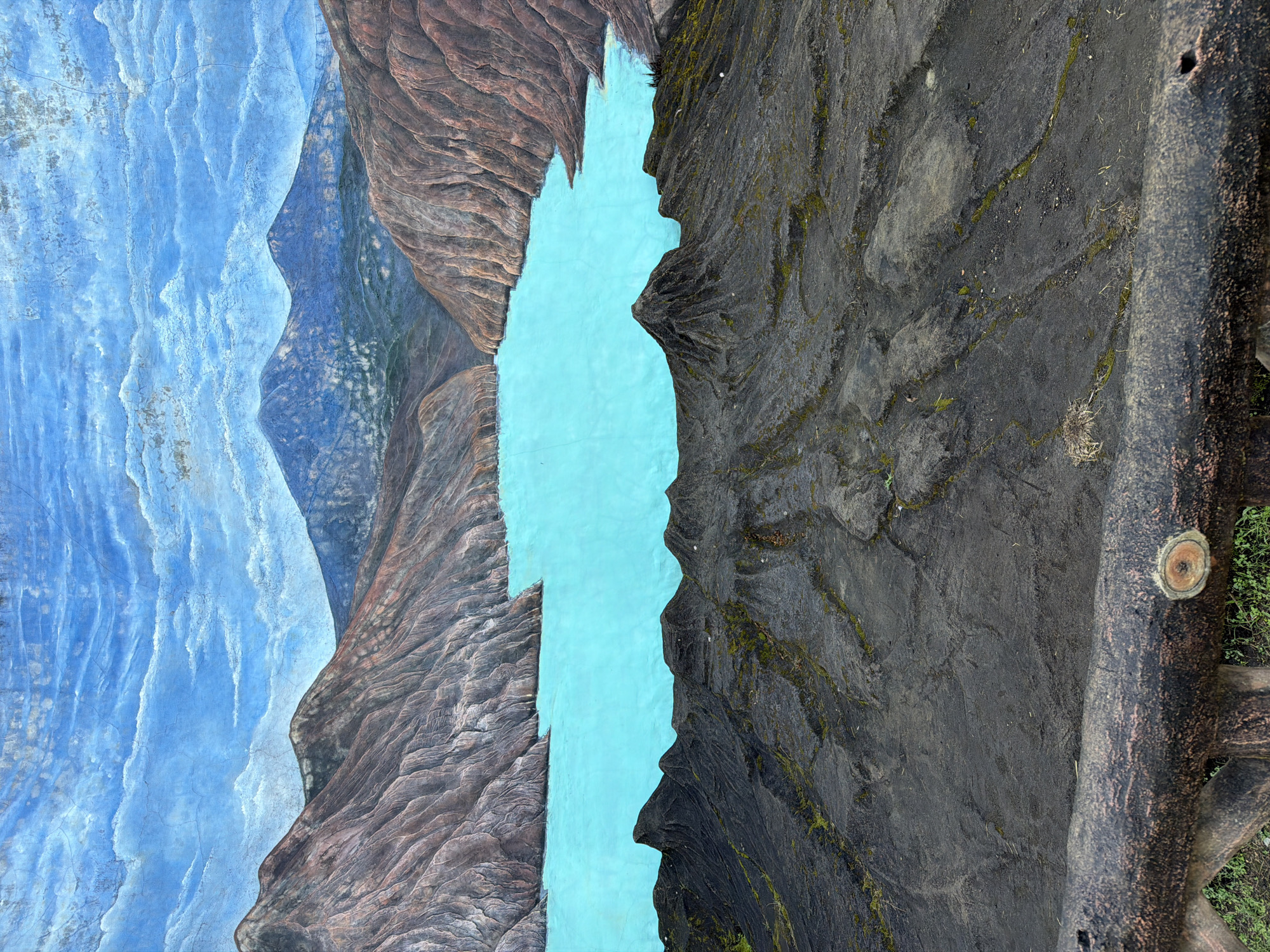The image depicts a mural of a landscape scene. On the left side, there's a depiction of blue, layered mountains blending into brown, rough-textured rock formations. A vertical turquoise-colored area separates the two sides. The right side features a darker, weathered rock formation. The mural is painted on what appears to be a textured surface, possibly concrete or a similar material. In the bottom right, a cut tree is present. The image seems to be taken outdoors in Unknown, Indonesia.