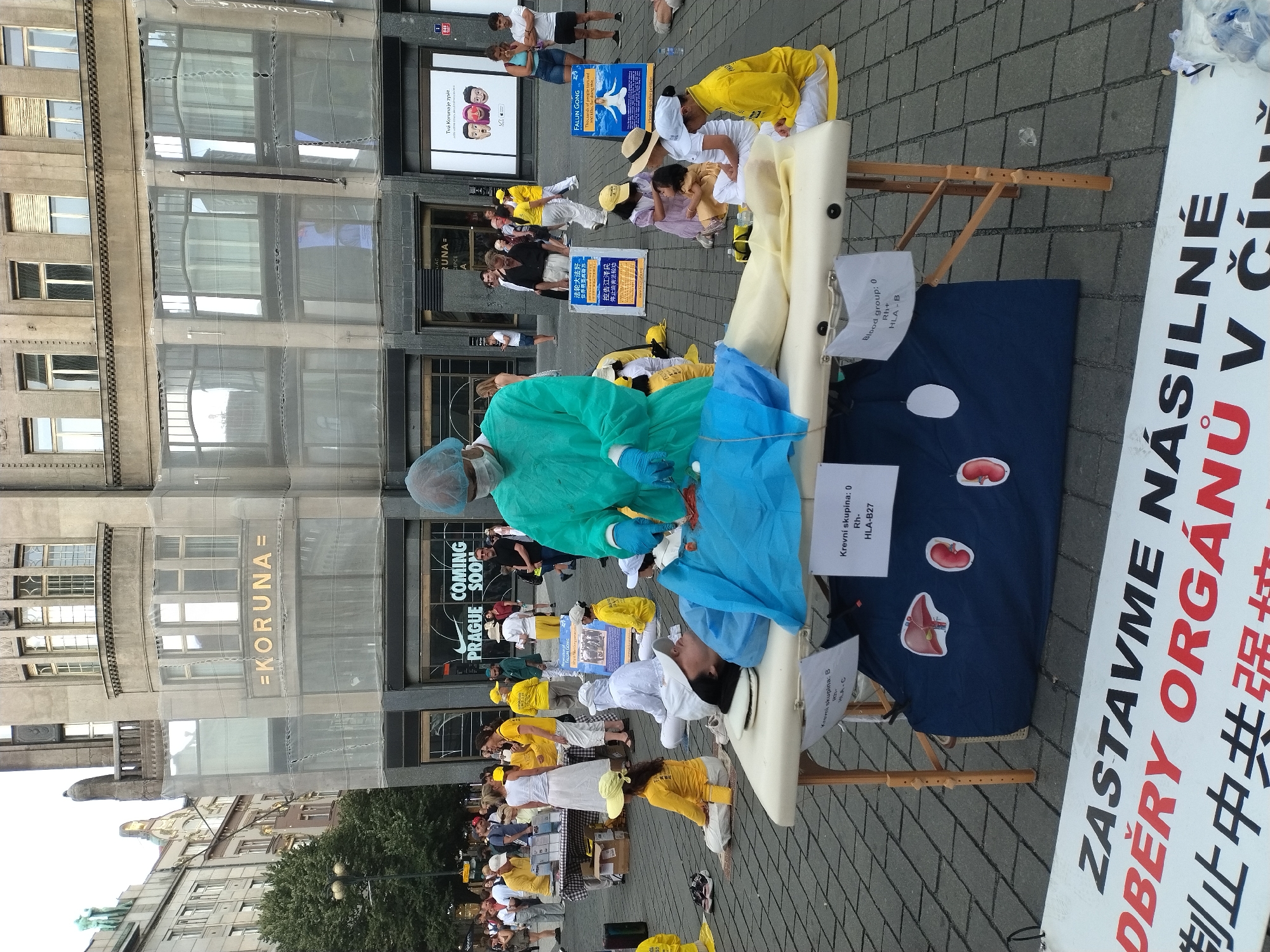 In Prague, Czechia, a street performance is taking place in front of a building with signage that includes "=KORUNA=". In the foreground, a table with a white sheet and a blue fabric spread over it represents a surgical operating table. A person in a teal surgical gown, mask, and gloves is simulating an organ transplant on a mannequin lying on the table. A dark blue fabric with images of a liver and kidneys is placed under the mannequin. There are signs with text in Czech and Chinese. Several individuals, some dressed in yellow, are seated on the ground around the operating table, appearing to be part of the protest or demonstration. In the background, a crowd of onlookers is present, and shops are visible, including one with a "COMING SOON" sign. The scene is set on a cobblestone street.