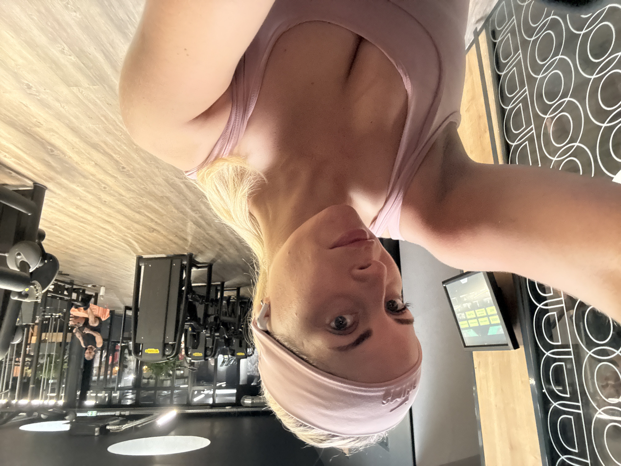A selfie of a blonde woman with a pink headband and a pink tank top is taken in what appears to be a gym. In the background, there are rows of gym equipment. Other people can be seen in the distance. The gym has large windows and a patterned design on the wall. The location is not discernible as being in Paris, France.