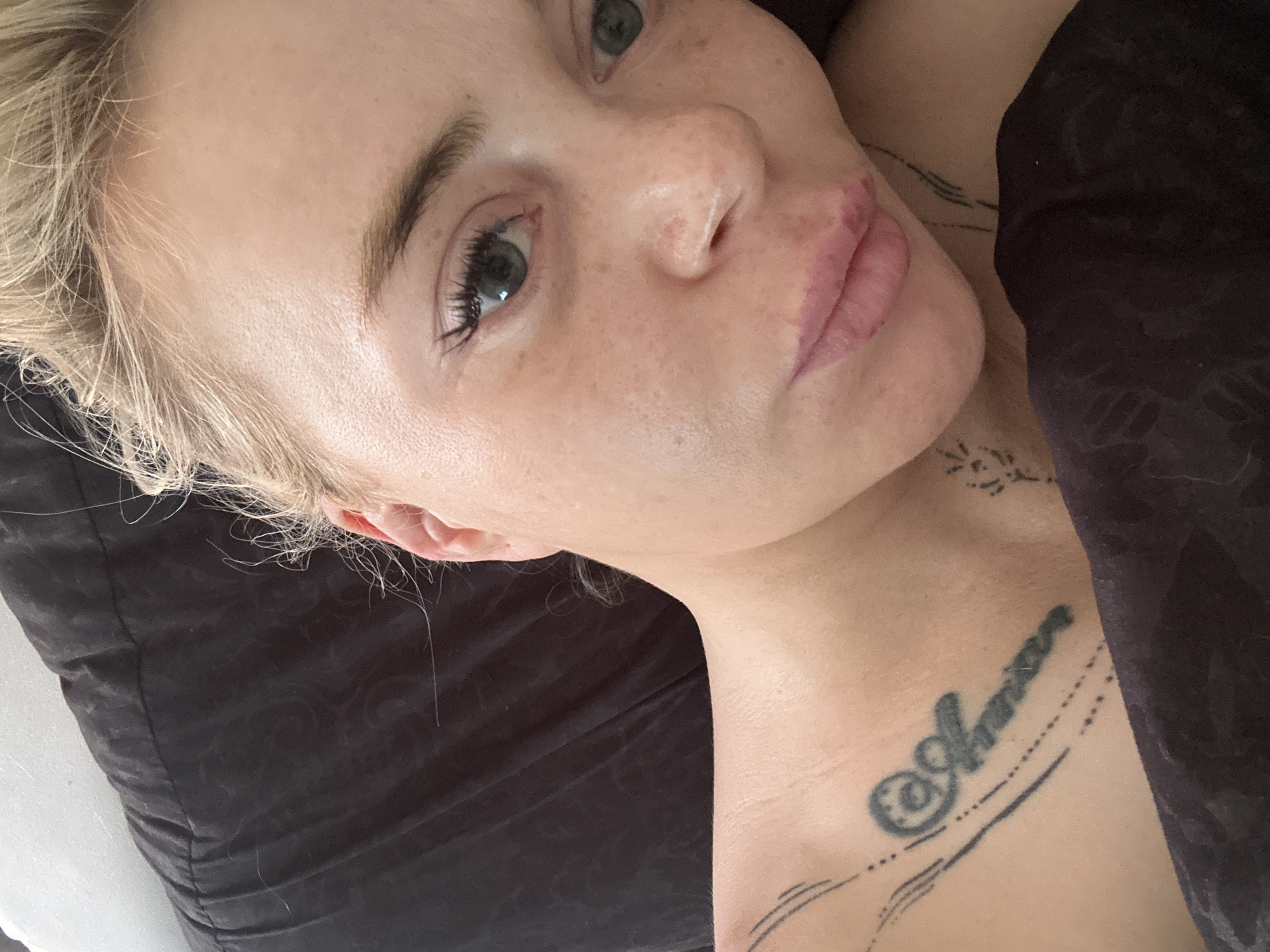 A close-up, selfie-style photograph shows a woman lying in bed, with her head resting on a dark pillow. She has blonde hair pulled back and is looking directly at the camera. Her face is tilted slightly, and she is wearing minimal makeup. There are several tattoos visible on her left shoulder and collarbone. The background consists of the dark pillow and a hint of white bedding. The setting is a bedroom, with no specific city or location identifiable.