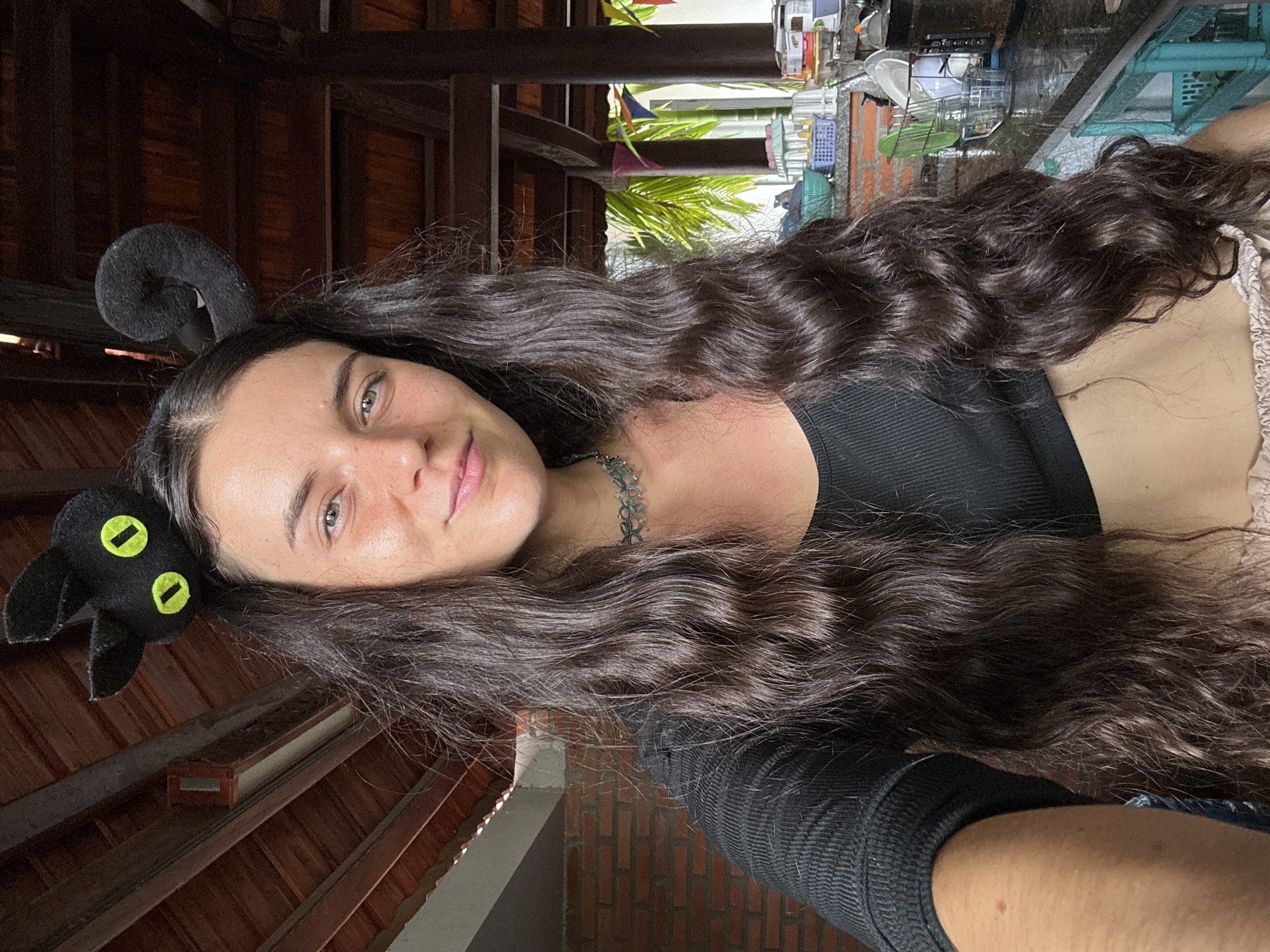 The image displays a close-up selfie of a young woman, presented rotated 90 degrees counter-clockwise. She has long, voluminous dark brown hair with natural waves, cascading over her shoulder and chest. Her face is slightly tilted, revealing clear, light-colored eyes and a gentle, relaxed smile. Freckles are visible across her nose and cheeks. She wears a distinctive black headband; on her right side (as viewed in the rotated image), there is a coiled, fuzzy black ear-like shape, while on her left side, a black felt cat's head with two bright yellow oval eyes is prominently featured. She is dressed in a black ribbed tank top and a dark, intricately designed choker necklace around her throat. A lighter, possibly cream-colored, garment is visible on her lower right side.
The background suggests an open-air yet covered space, potentially a porch or a section of a traditional building, aligning with the Mũi Né, Vietnam location context. Prominent wooden beams and slats form the ceiling and structural components. Beyond the immediate structure, lush green palm foliage is visible, indicating a tropical environment. Further in the background, a collection of household items, including baskets and drying dishes, along with a partial brick wall, suggests a local, residential setting. The scene is illuminated by bright natural daylight, implying the photo was taken during the day. No visible text is present in the image.