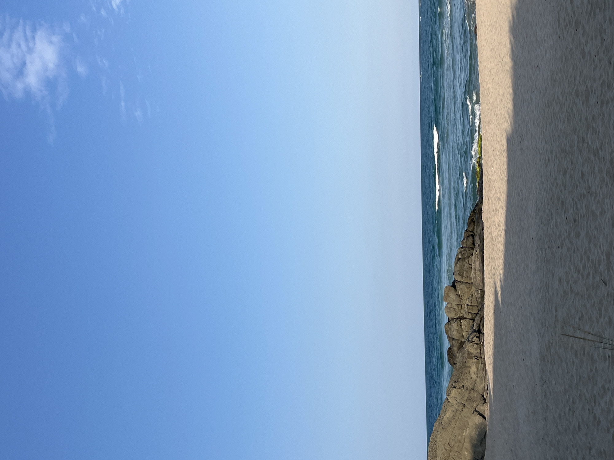 The image depicts a serene beach scene in Ahtopol, Bulgaria. A clear, bright blue sky dominates the upper portion of the frame. Beneath it, the deep blue sea meets the shore with gentle waves. A line of large, dark rocks juts out into the water from the left side of the image, providing a rugged contrast to the soft, light-colored sand of the beach in the foreground. The sand is smooth and shows subtle undulations from wind or water. The overall impression is one of tranquility and natural beauty, characteristic of Ahtopol's coastline. There are no people visible in the image, emphasizing the peacefulness of the location.