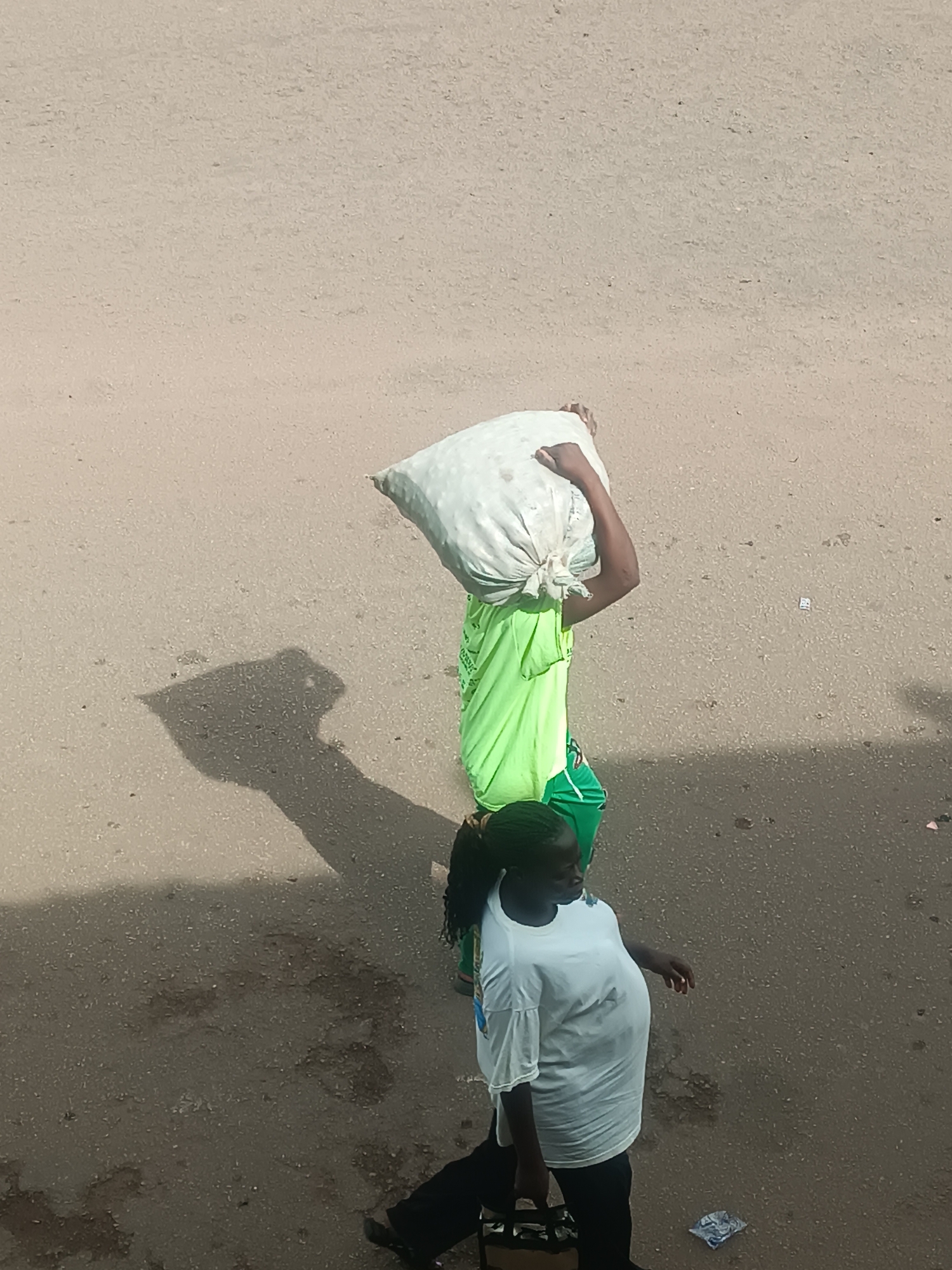 The image depicts an aerial view of two individuals on a road in Bukuru, Nigeria. One person carries a large white sack on their head, wearing a bright green garment. A second individual, in a white shirt and dark pants, walks alongside them. The road appears to be made of asphalt, with patches of dirt and scattered debris. A shadow is cast by the figure carrying the sack, indicating sunlight.