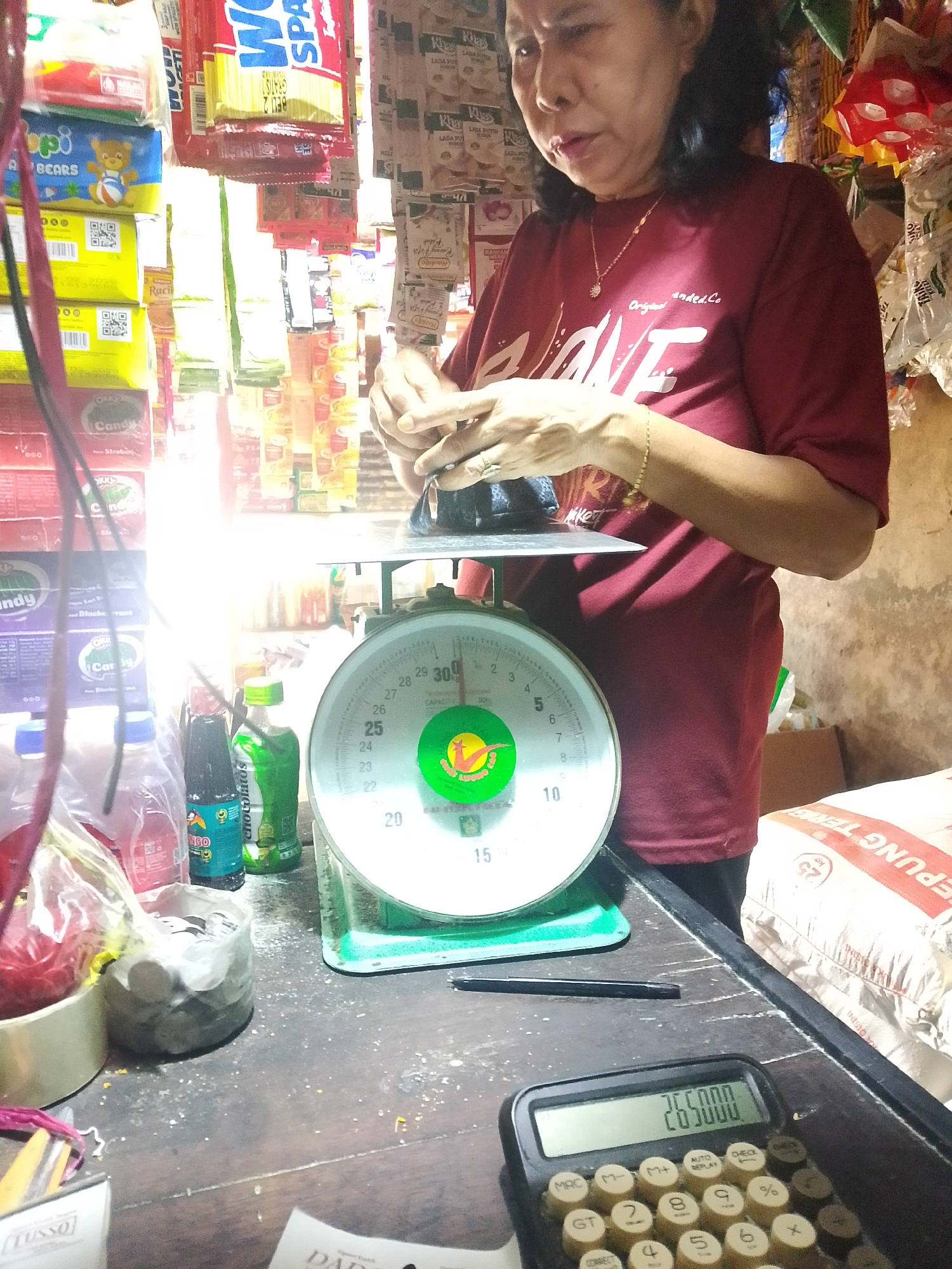 The image depicts a retail environment, likely a small shop or market stall, in Palembang, Indonesia. A woman, appearing to be the shopkeeper, is in the process of weighing an item on a green mechanical scale. She is wearing a red t-shirt with a graphic design and a gold necklace. Various products, including packaged snacks, candies, and drinks, are visible in the background, stacked on shelves and hanging from displays. A calculator showing "265000" is in the foreground, along with other miscellaneous items on a wooden counter. The lighting suggests an indoor setting, possibly with natural light entering from the side, creating bright highlights and shadows. The overall atmosphere is that of a busy, everyday transaction.