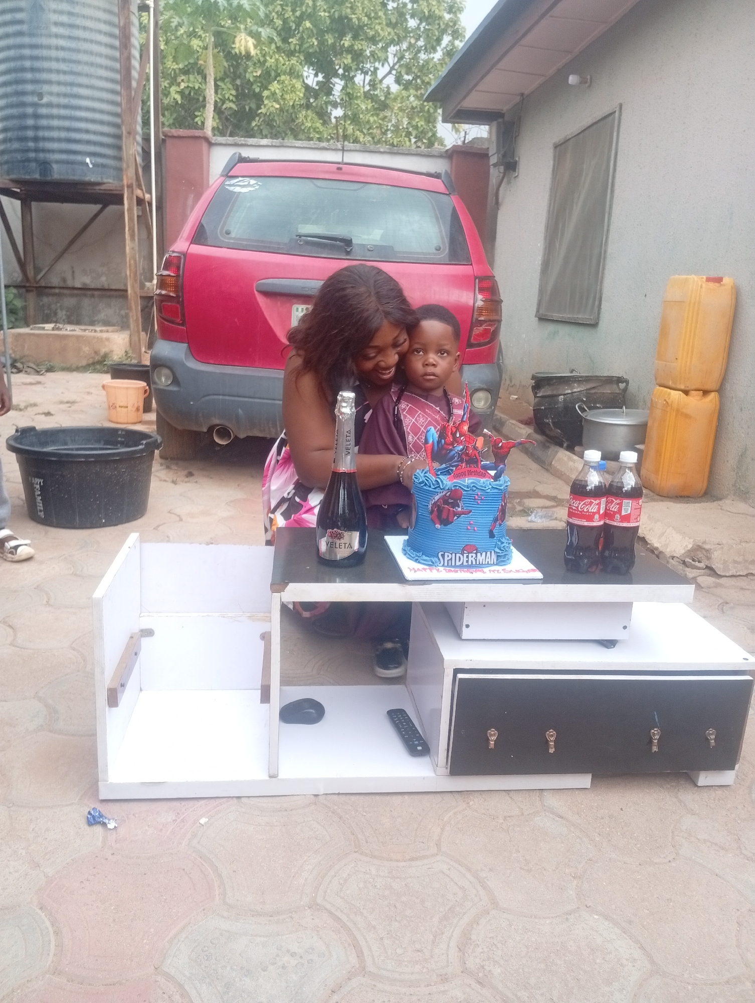This image captures an outdoor birthday celebration in Sabon Tasha, Nigeria, on a bright day. A smiling woman embraces a young boy, likely the birthday celebrant, who looks directly at the camera. They are positioned behind a modern, multi-level white and black table.
On the table, a blue Spiderman-themed birthday cake is prominent, inscribed with "Happy Birthday" and "SPIDERMAN." Beside the cake are two bottles of Coca-Cola and a bottle of Veleta sparkling wine. The woman wears a patterned dress, and the boy is dressed in a patterned top. Their expressions convey happiness and warmth.
The setting appears to be a residential area with paved interlocking concrete blocks. In the background, a red SUV is parked, and a large, ribbed black water storage tank stands on a metal frame. Various other items are visible: a black bucket labeled "HAPPY FAMILY" on the ground, a brown bucket, two yellow jerrycans stacked against a building wall, and a cooking pot. The small table also features black drawers with distinctive gold key-shaped handles. A TV remote control and a computer mouse are also visible on the table's lower shelves. The scene suggests a joyful, casual gathering.