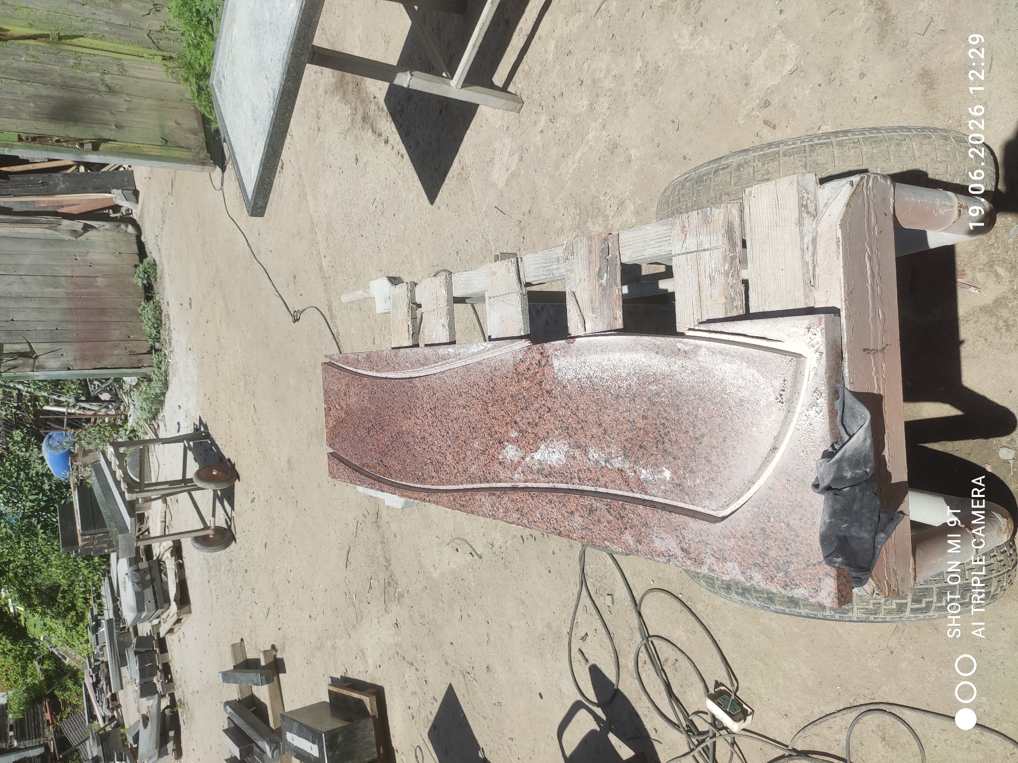 In an outdoor area with a sandy, dirt ground, a reddish-brown granite slab with a curved edge is positioned on a wheeled wooden platform. The slab appears to be undergoing shaping or finishing, as indicated by a white powdery residue along the cut edge. Various tools and equipment, including extension cords and an electrical outlet box, are present near the slab. In the background, there are stacks of stone slabs, a wooden fence, and vegetation. The scene suggests a workshop or yard where stone processing takes place. The watermark "SHOT ON MI 9T AI TRIPLE CAMERA 19.06.2026 12:29" indicates the image was captured on June 19, 2026, at 12:29 PM.