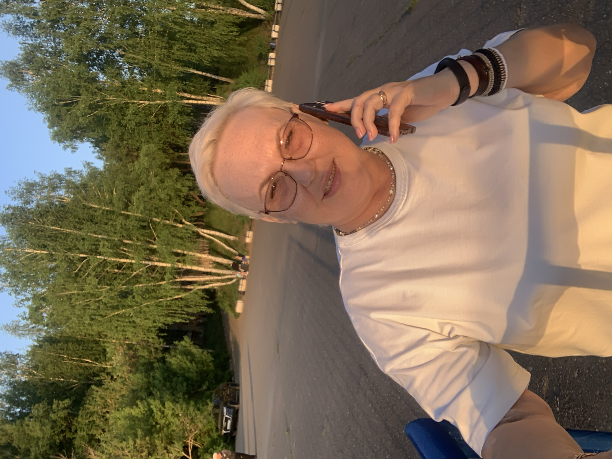 A fair-skinned woman with short blonde hair and glasses is holding a phone to her ear in the city of Chaikovsky, Russia. She is wearing a white t-shirt, a necklace, and bracelets. A paved road and trees are visible in the background.