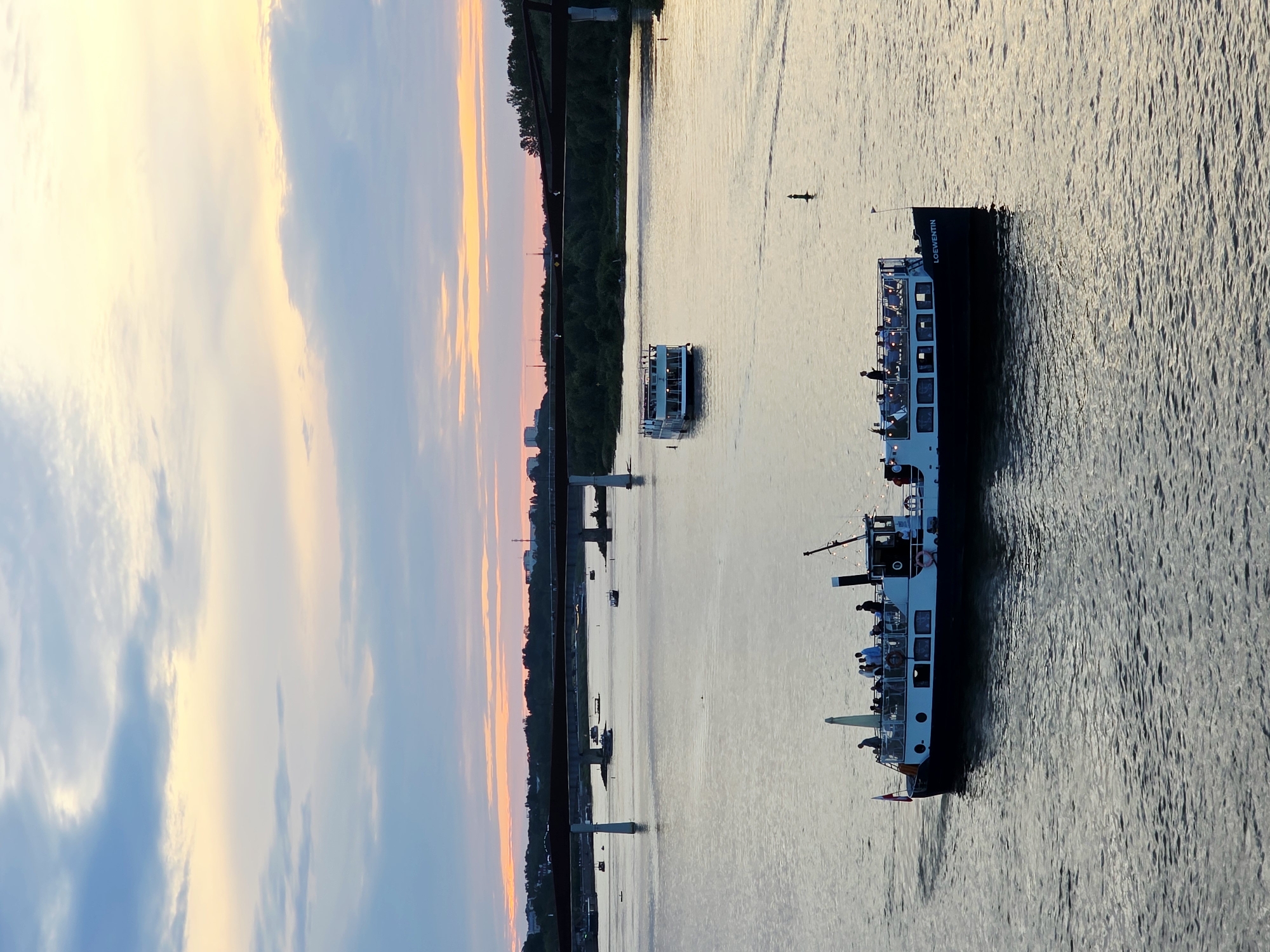 This image captures a serene sunset over the Vistula River in Warsaw, Poland. The sky is painted with hues of orange, yellow, and blue, with scattered clouds. A large bridge spans the river in the background, and several boats are visible on the water. In the foreground, a passenger boat named "LOEWENTIN" is cruising, with people visible on its deck, enjoying the view. Another, smaller boat is seen further down the river. The water reflects the colors of the sky, creating a tranquil and picturesque scene. The city's skyline is faintly visible in the distance.