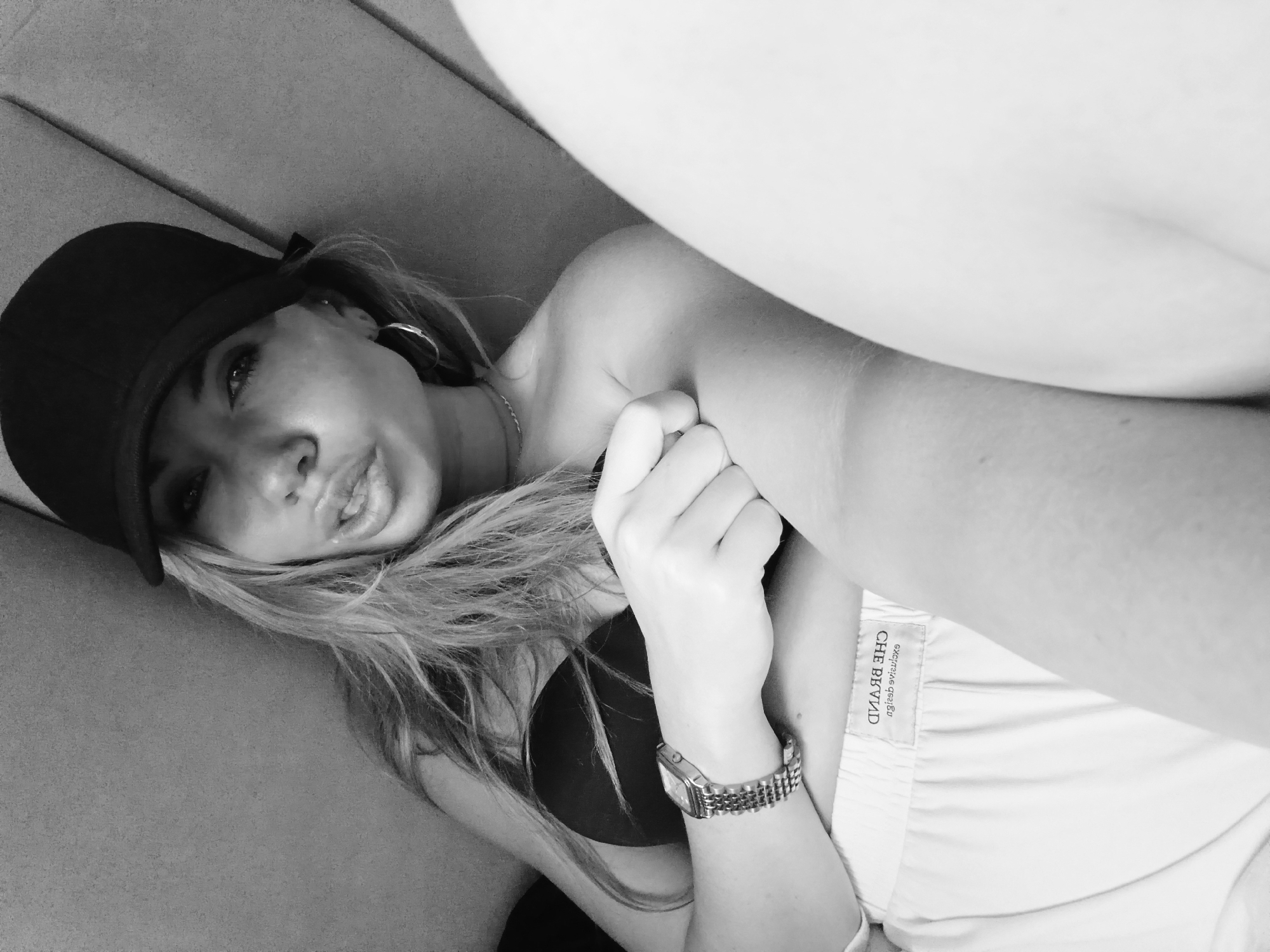 A black and white close-up of a woman is presented in a low-angle shot. She wears a black hat, a strapless top, and a watch on her left wrist. Her hair is light-colored. She is inside what appears to be a car or a location with a cushioned interior, suggested by the grey surface at the top left of the image. There is no discernible evidence within the image to suggest a location in Votkinsk, Russia.