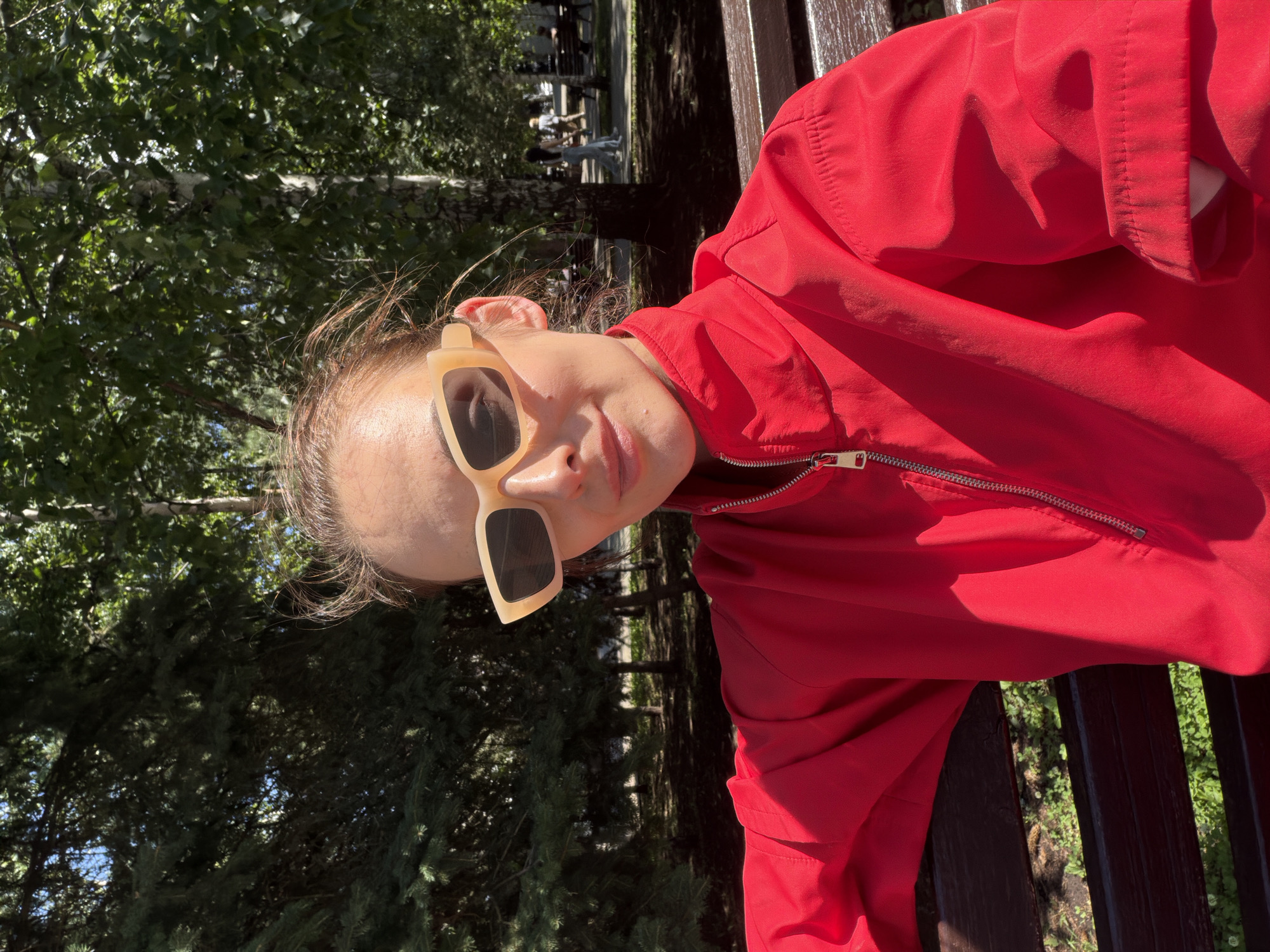 A young woman with light brown hair pulled back is sitting on a park bench. She is wearing oversized light-colored sunglasses and a bright red jacket. The background shows a park with trees and other people walking in the distance. The lighting suggests it is daytime. The location is the Salavat Yulaev monument park in Ufa, Russia.