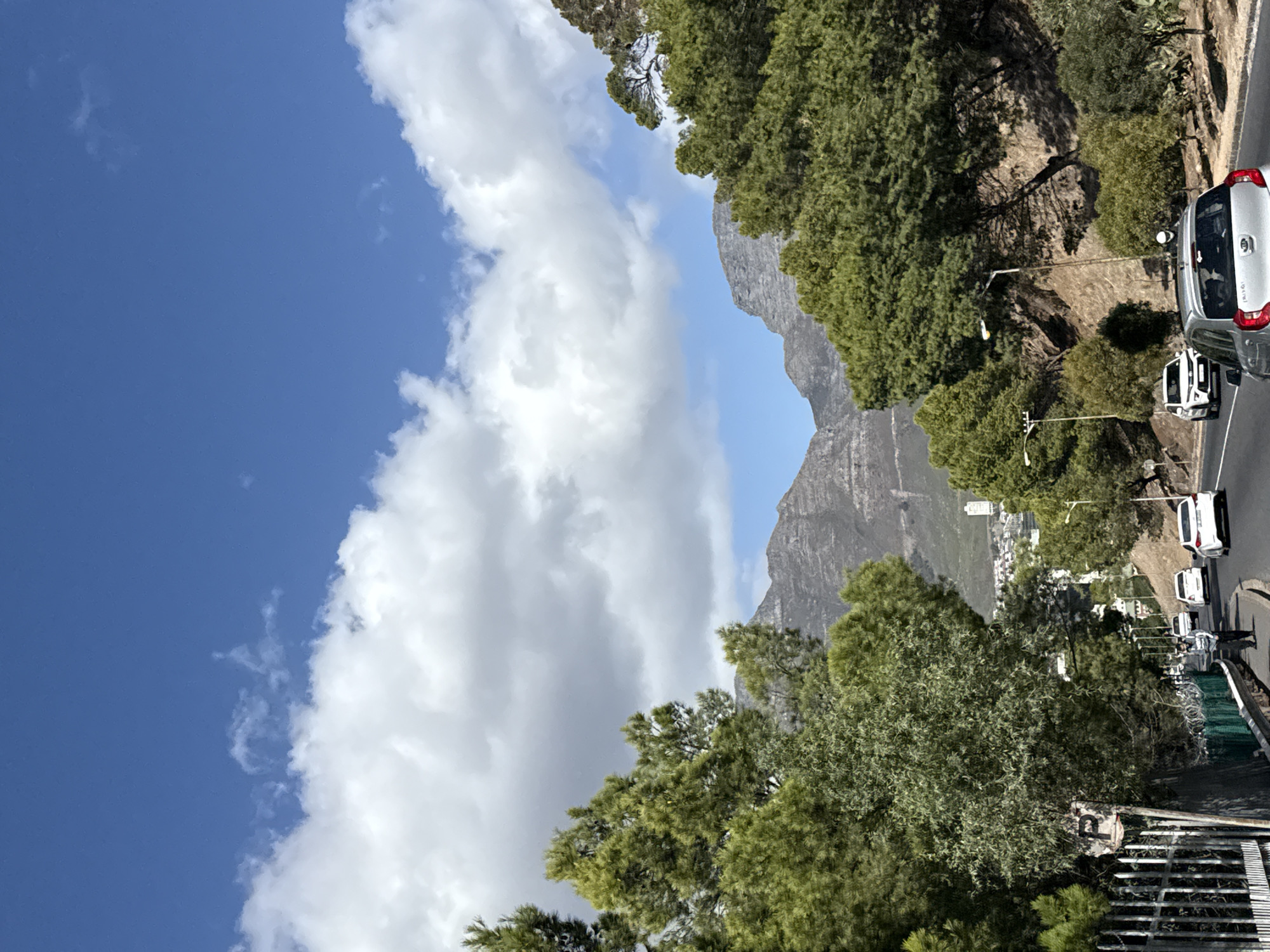 The image shows a view of Cape Town, South Africa, featuring Table Mountain in the background. The sky is blue with scattered white clouds. A road with several parked and moving cars is visible, with vegetation on the side. Street lights are present along the road. The viewpoint appears to be elevated, providing a scenic perspective.