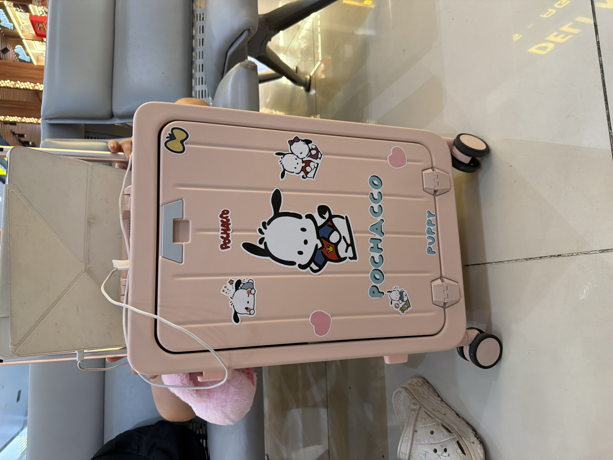 A pink suitcase is positioned in the foreground of the image, resting on the floor and partially obscuring a white clog on the left. The suitcase features several stickers, including characters from Sanrio's Pochacco franchise, and the word "POCHACCO" emblazoned in teal lettering. A white charging cable is connected to a tablet or laptop resting on top of the suitcase. The background shows seating, likely in a waiting area, and illuminated ceiling fixtures, suggesting an indoor public space such as an airport terminal or a shopping mall. The floor is tiled with a grid pattern. No individuals are clearly depicted, though a glimpse of a person's arm and shoulder is visible on the left, and another person's hand is seen near the tablet. The scene takes place in a well-lit environment.