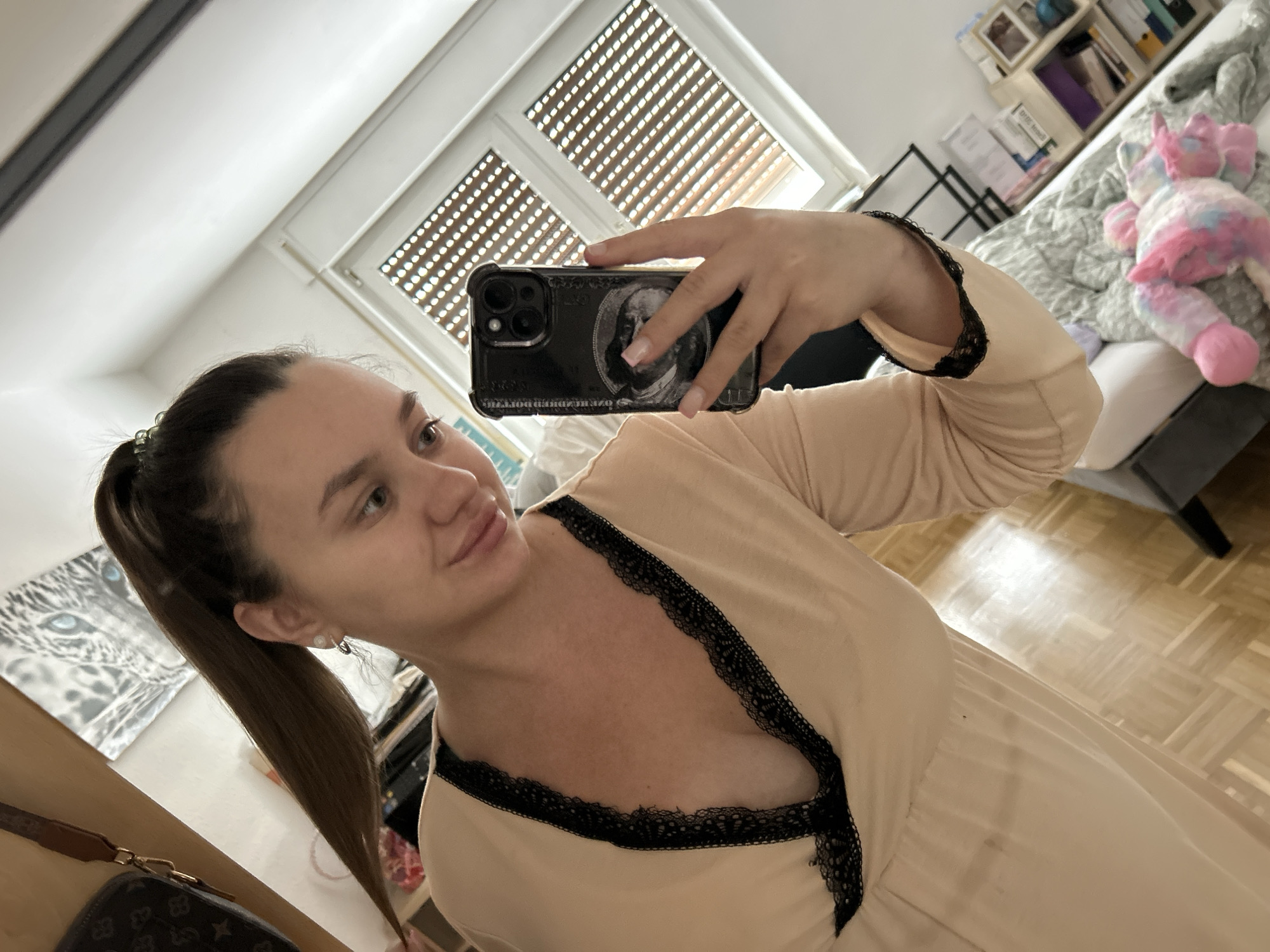 A woman with her hair in a high ponytail takes a selfie in a room. She is wearing a pale peach-colored dress with black lace trim along the neckline and sleeves. The dress has a deep V-neck. She is holding a black smartphone in her right hand. In the background, there is a window with horizontal blinds, a shelf unit filled with books and decorative items, and a bed with a colorful plush toy. The floor is made of light-colored wood. The overall impression is of a domestic setting, likely a bedroom or living area. This image is not sufficient to determine the location in the city of Waldbröl, Germany.