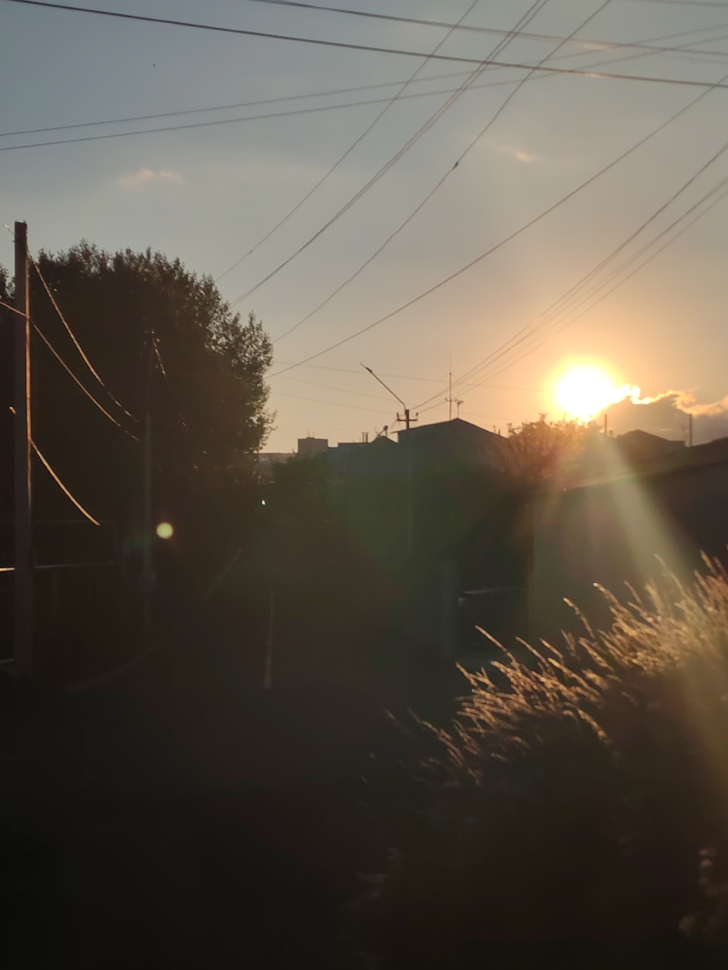 The image depicts a sunset in Sevan, Armenia. The sun, a bright orb, is setting in the sky, casting a warm glow. In the foreground, there is a silhouette of a building, possibly a residential structure, with a few trees and power lines visible. The scene is dimly lit, with the setting sun creating a lens flare effect. The overall atmosphere is serene and peaceful, capturing the natural beauty of the sunset over the city.