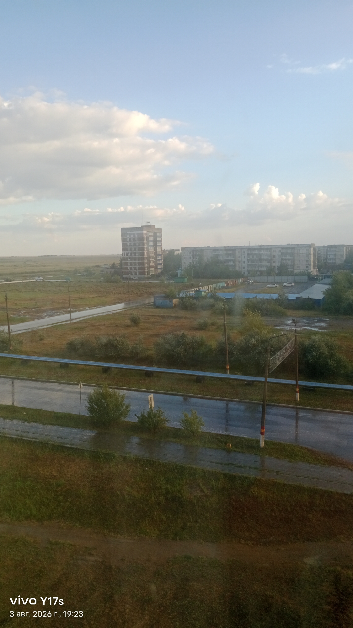 The image depicts an outdoor scene in the city of Rudny, Kazakhstan, likely during or after rainfall. In the foreground, a paved road with standing water and reflections is visible, bordered by a grassy embankment and vegetation. A blue railing runs along the edge of the embankment. Further back, more greenery and trees are present, along with a paved pathway. Several utility poles are scattered throughout the scene. In the middle ground, there are low-lying buildings, possibly garages or sheds, and a larger, multi-story brick building in the background. Beyond this, a flat, open field stretches to the horizon, with what appear to be residential buildings in the distance under a partly cloudy sky. The timestamp in the lower left corner indicates the image was captured on August 3, 2026, at 19:23. No people are visible in the frame.