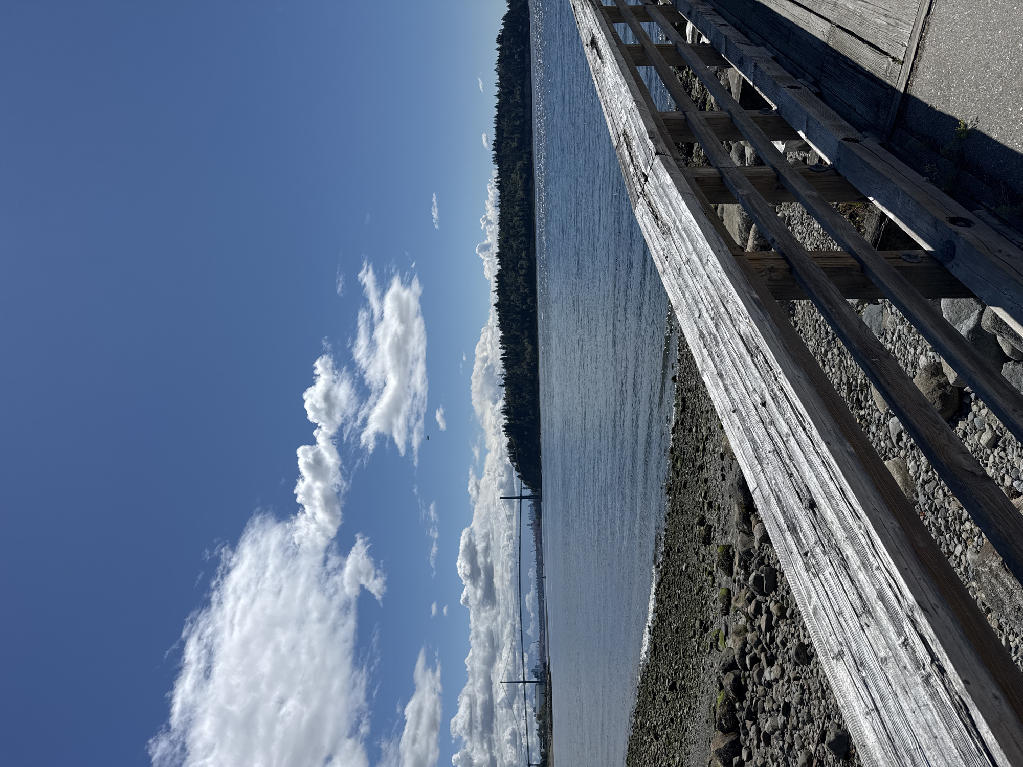 The image shows a view from a wooden pier looking out at the water and a distant bridge. The pier railing is in the foreground, made of weathered, light-colored wood. The water is calm and blue, with small waves lapping at a rocky shore. In the distance, a large suspension bridge spans the water, and behind it, a tree-covered island or peninsula is visible. The sky is a clear blue with scattered white clouds. The scene is set in West Vancouver, Canada, likely near the Lions Gate Bridge, given the presence of the distinctive suspension bridge. There are no people visible in the image.