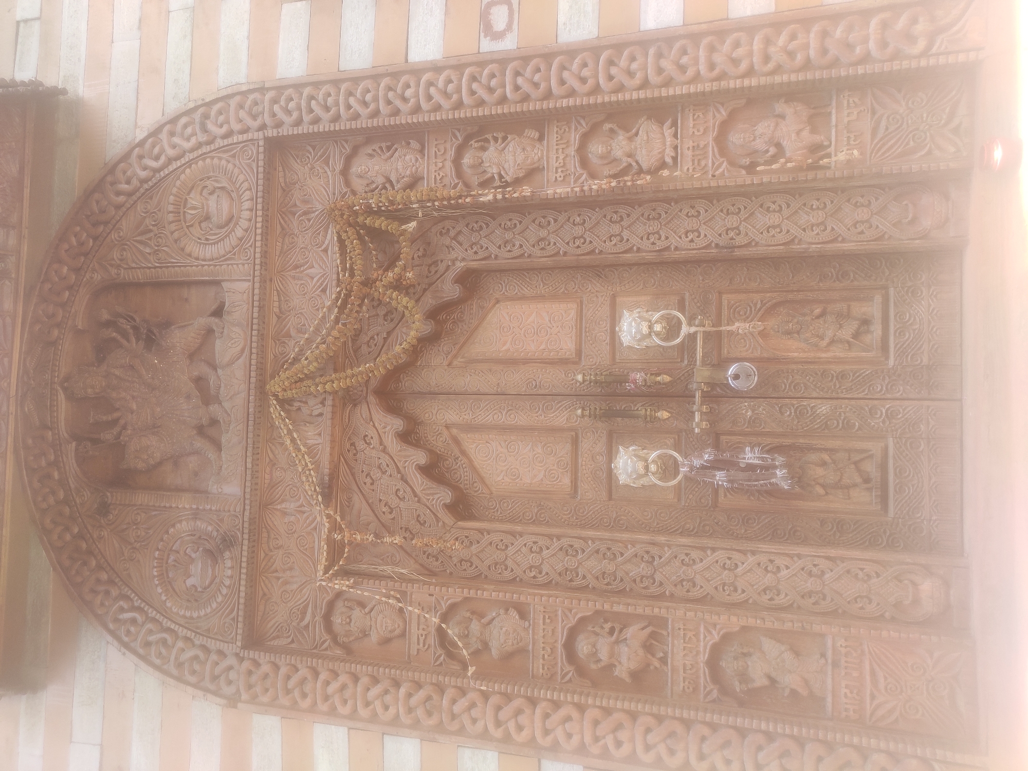 This is a frontal, eye-level, slightly hazy, medium shot of a large, intricately carved wooden double door, likely part of a temple in Naggar, India. The door is adorned with numerous carvings, including figures of deities, floral patterns, and decorative borders. A string of dried flowers or beads hangs across the upper portion of the door. The door has two ornate lion-head knockers and a central locking mechanism with a padlock. The surrounding walls are made of stone or brick with horizontal banding. A small, lit candle is visible at the bottom right of the frame. The overall impression is of a sacred entrance with rich cultural and religious significance.