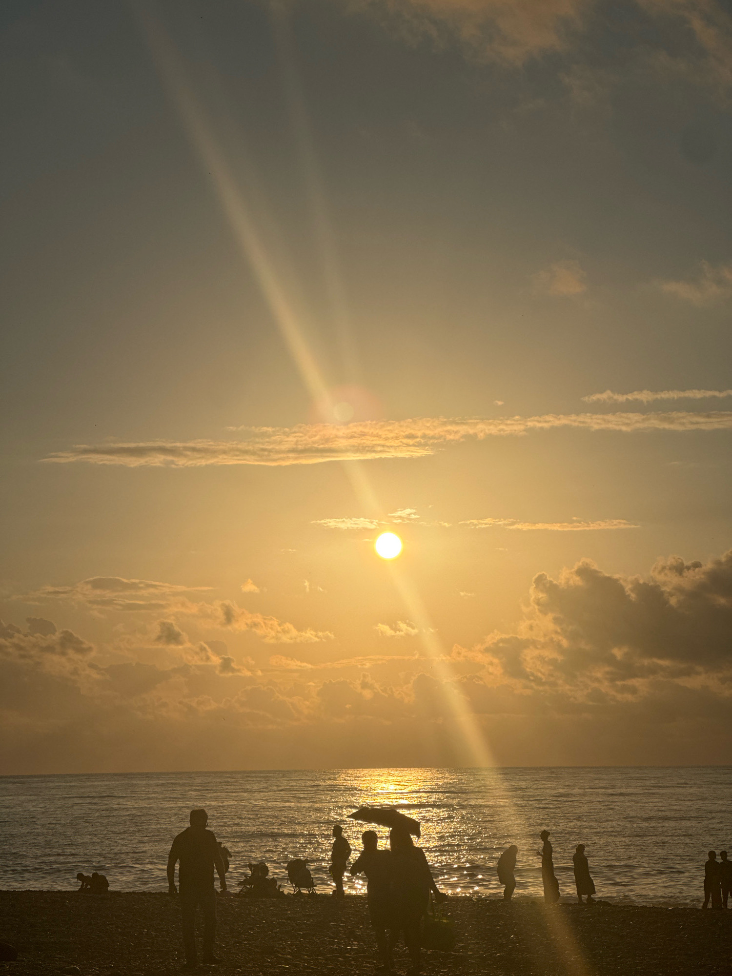 The image depicts a sunset on a pebble beach, likely in Batumi, Georgia, given the presence of palm trees (though not clearly visible in this specific frame) and the characteristic Black Sea coastline. The sun is setting over the water, casting a warm, golden glow across the sky and sea. The sun's rays are visible, fanning out from the horizon and illuminating scattered clouds. Silhouetted figures of people are scattered along the shoreline, some walking, others standing or sitting. Some individuals appear to be near the water's edge, while others are further back on the beach. The sea is calm with gentle waves lapping at the shore. The overall atmosphere is serene, with a sense of peaceful evening activity.
