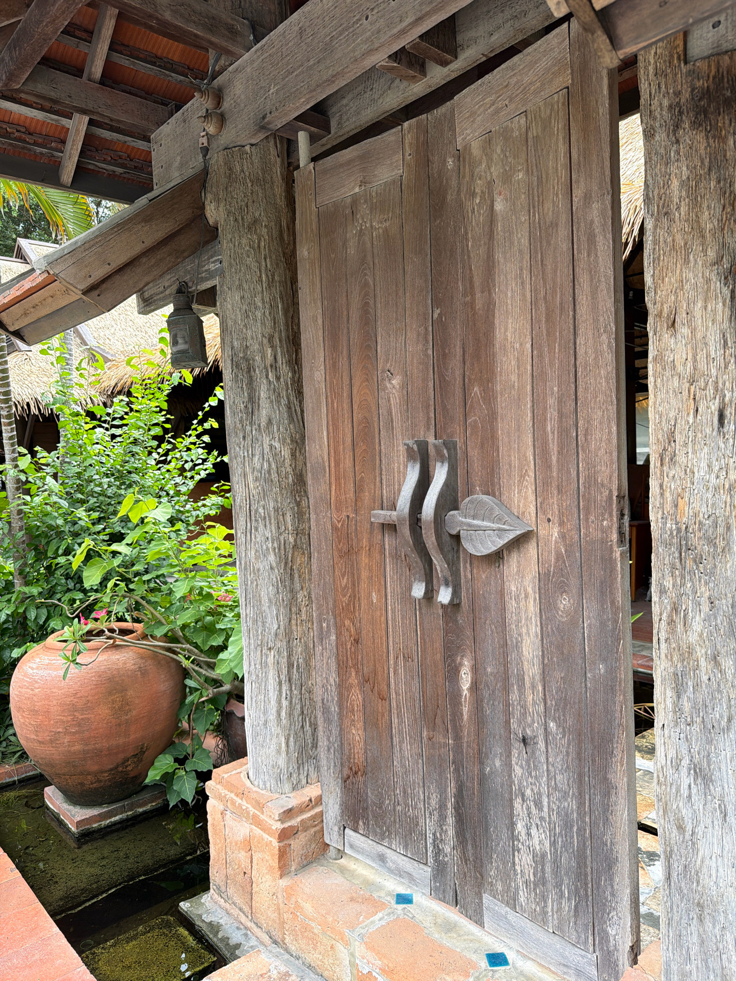 An open wooden door reveals a glimpse into a building with a thatched roof in Ko Samui District, Thailand. The door, constructed from vertical wooden planks, features a decorative wooden handle shaped like a leaf. To the left of the door, a large, rustic clay pot sits beside a small water feature. Lush green foliage surrounds the pot, and a straw-thatched roof is visible in the background, suggesting an outdoor or semi-outdoor setting. The scene is framed by rough-hewn wooden posts and beams, contributing to a natural and rustic aesthetic.