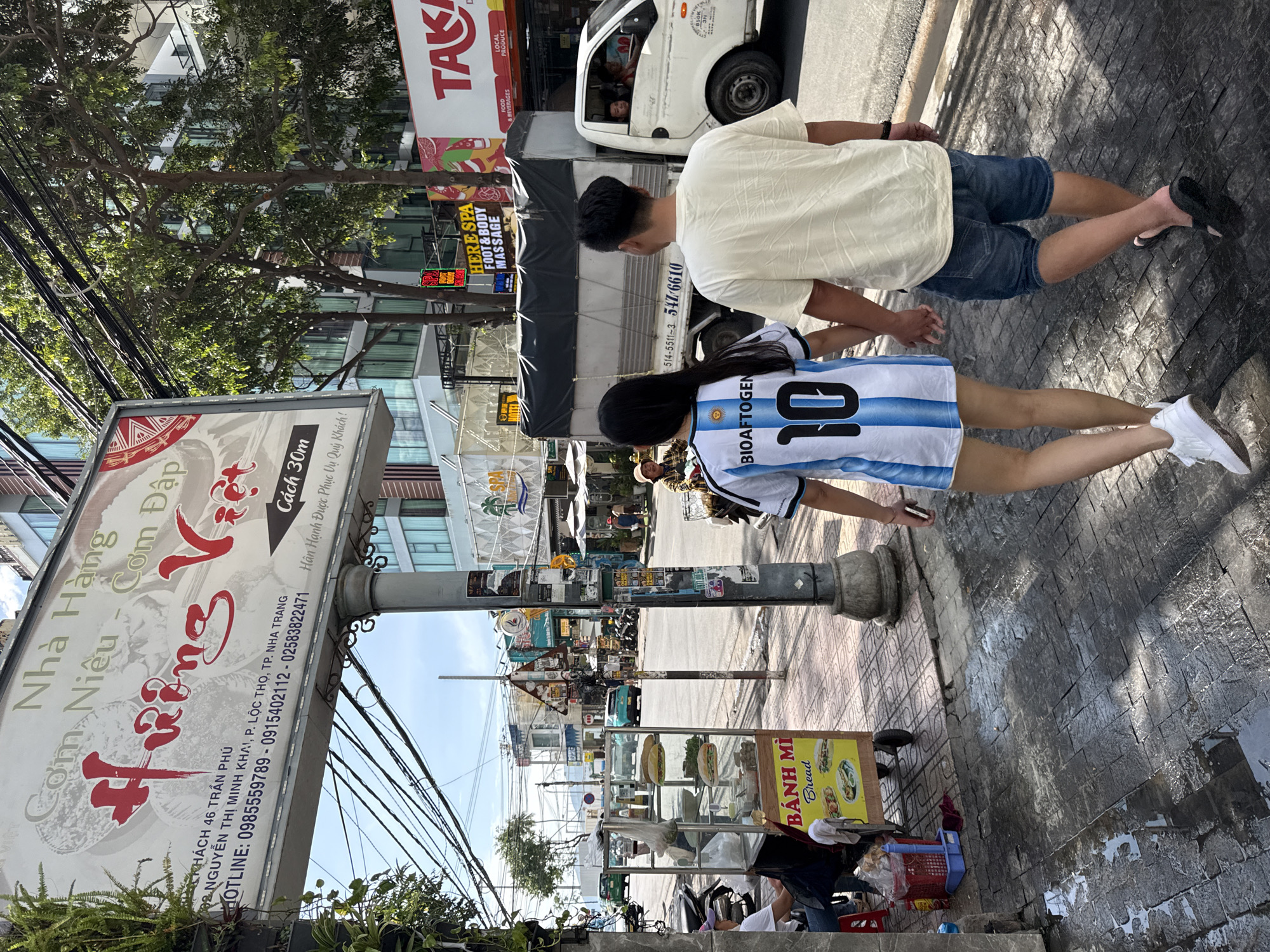 In Nha Trang, Vietnam, two individuals, a woman and a man, walk hand-in-hand down a city street. The woman wears a white and blue striped soccer jersey with the number 10 on the back and "BIOAFTOGEN" printed above it, paired with white sneakers. The man is dressed in a cream-colored t-shirt and denim shorts, wearing black flip-flops. They are walking away from the camera on a wet, dark-tiled sidewalk, which reflects the sunlight.
The background features a street scene with various businesses and vehicles. A food cart selling "BÁNH MÌ" (Bread) is visible on the left, with sandwiches displayed in a glass case. A large sign for a restaurant named "Nhà Hàng Hương Việt" is prominent above. Further down the street, buildings with multiple stories line the road, some with advertisements and awnings. A white truck with a tarp-covered bed is parked on the right side of the street, and a light blue car is driving past on the road. The environment suggests a warm, sunny day, with some wetness on the pavement indicating recent rain or cleaning. Overhead, electrical wires crisscross the sky.