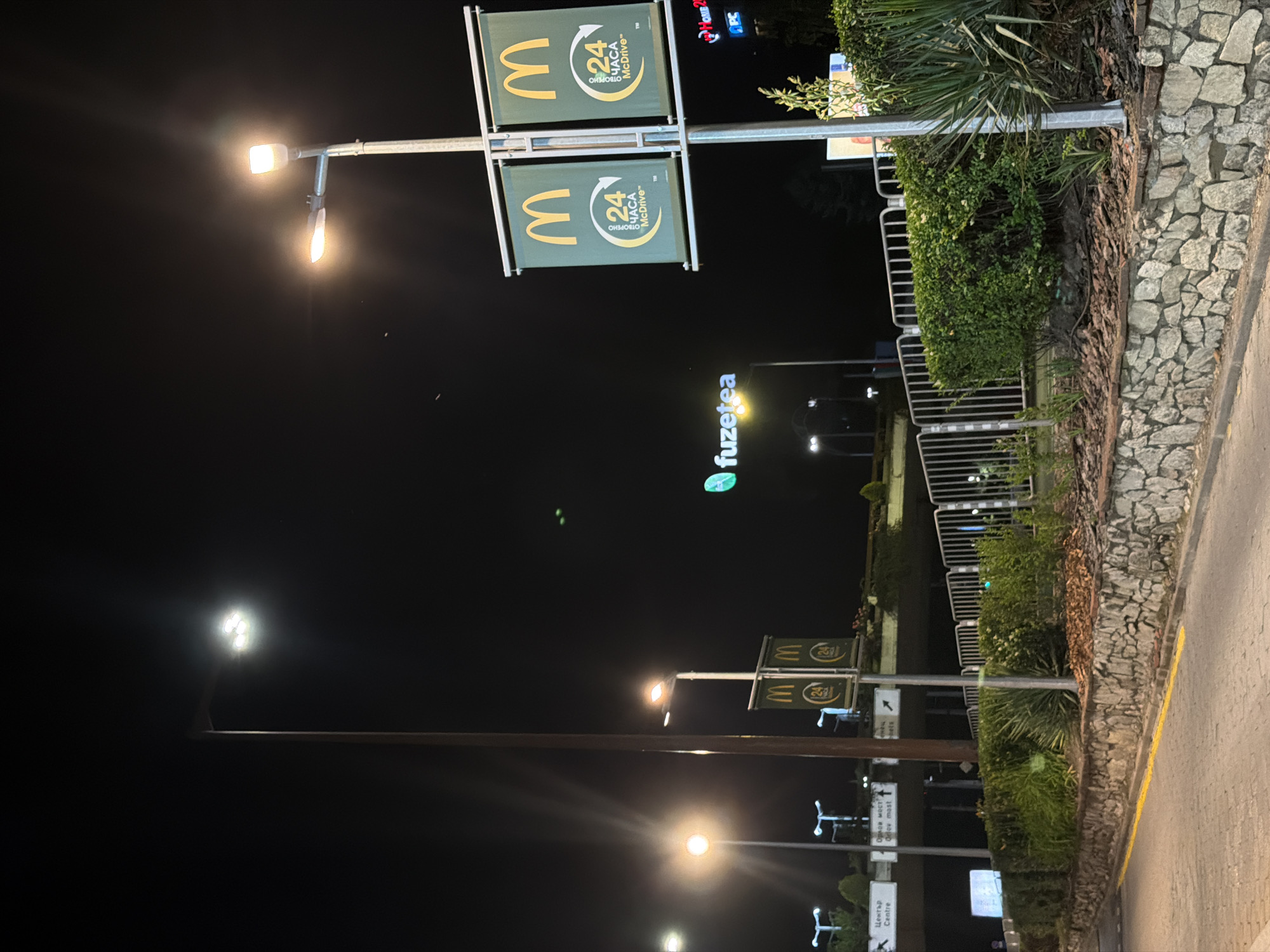 The image depicts a nighttime scene in Sofia, Bulgaria, likely near a commercial area. In the foreground, a stone-lined embankment borders a paved road with a yellow line marking its edge. Lush green hedges and some spiky plants are visible on the embankment.
Dominating the middle ground are two large banners featuring the McDonald's logo and "24 ЧАСА McDrive" (24 Hours McDrive), indicating a drive-thru service. These banners are mounted on a pole alongside a street lamp. Further back, there's a sign for "fuzetea" with its distinctive green logo illuminated.
Above the banners and fuzetea sign, an elevated structure, possibly a highway or overpass, is visible. Road signs in Bulgarian are also present on this structure. Several street lamps illuminate the scene, their light creating a bright, nighttime atmosphere. The background is dark, suggesting a clear night sky. No people are clearly visible in the image.