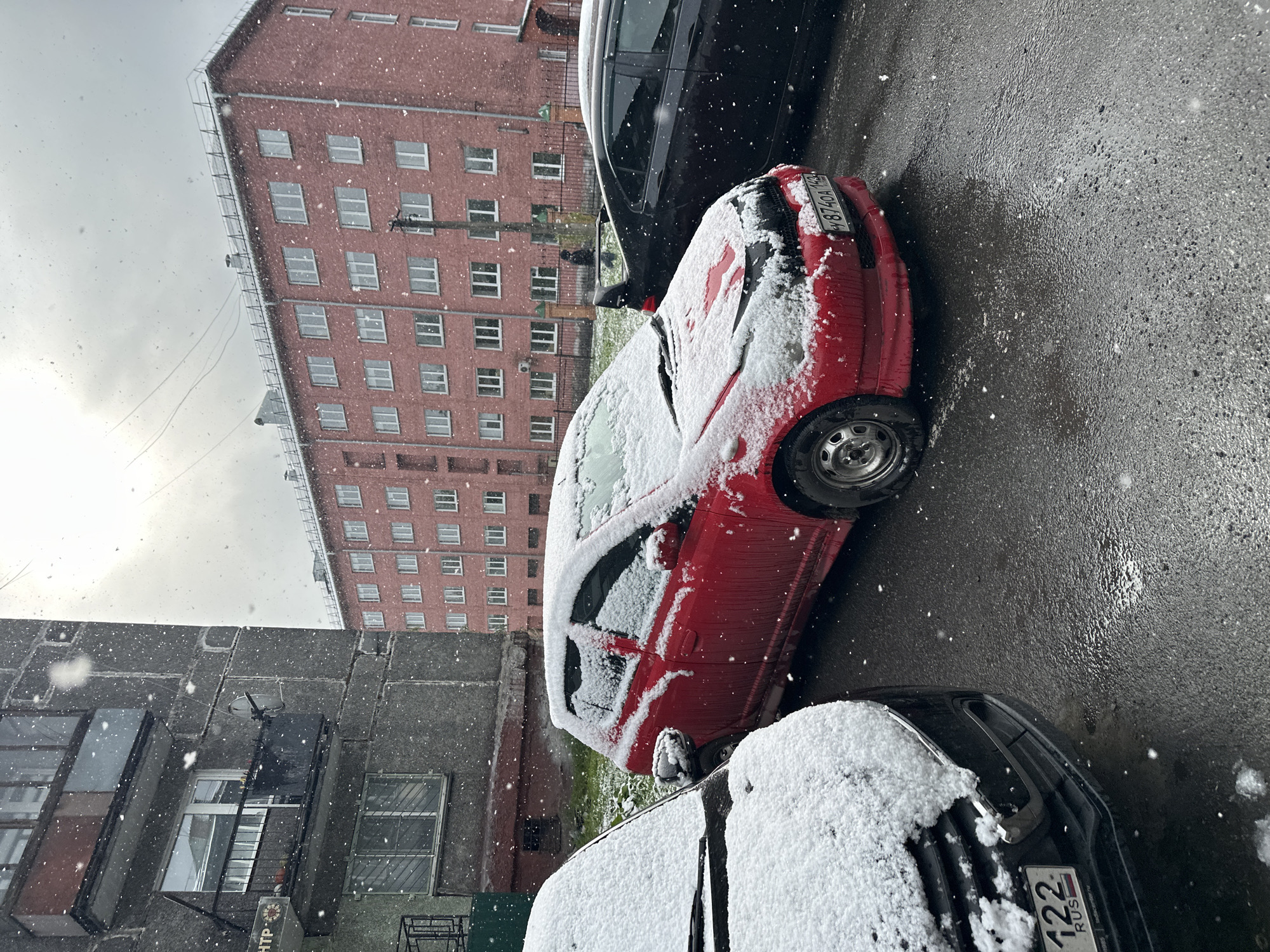 The image captures a snowy day in Novokuznetsk, Russia. A red car is parked alongside a black car, both partially covered in snow. The street is wet, suggesting melting snow or rain. Buildings are visible in the background, one a tall red-brick structure with numerous windows, and another a grey, older building. Snow is falling, and flakes are visible in the air. License plates on both vehicles are visible. The Russian city's name cannot be clearly distinguished from the license plates' region code. The scene is typical of winter weather in a Russian urban environment.