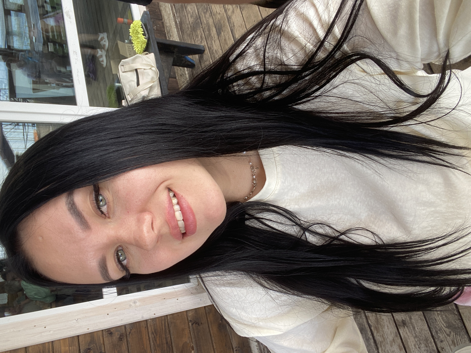 The image shows a medium shot of a young woman with long, dark hair, pale skin, and green eyes. She is smiling and looking at the camera. She is wearing a light-colored long-sleeved shirt and a delicate necklace. The background appears to be an outdoor setting, possibly a deck or patio area with wooden structures and a large window reflecting other people and an indoor environment. On a table in the background, a light-colored bag and a bright green object are visible. The overall scene suggests a casual outdoor gathering. Without further context, it is not possible to definitively place this image in the city of Chaikovsky, Russia.