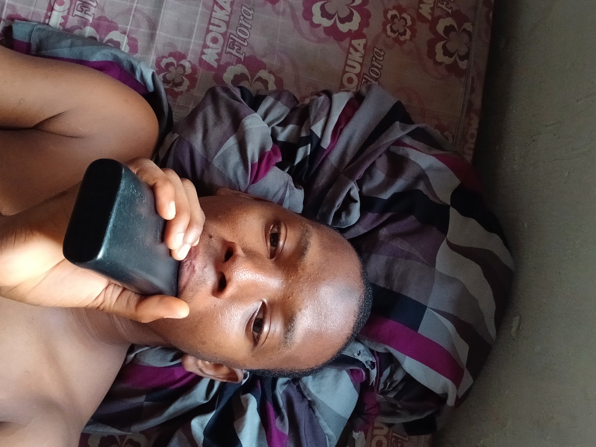 An African man lies on a bed with a floral-patterned mattress and a gray and purple blanket. He is holding a black rectangular object, possibly a cell phone, up to his ear. The scene appears to be indoors, with a wall visible in the background. The location is indicated to be Lagos, Nigeria, though specific landmarks or features confirming this are not visible in the image.