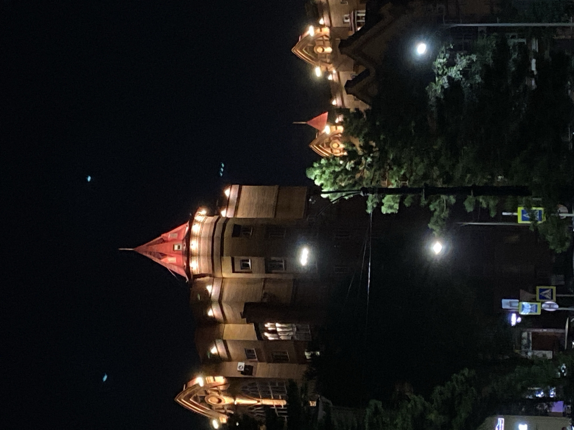 A night-time photograph captures an illuminated building with a distinct red-capped turret. The building appears to be made of brick and has multiple windows, some of which are lit. Adjacent to the main structure are other buildings with illuminated architectural details. In the foreground, trees are visible, obscuring the lower portion of the buildings. Streetlights and traffic signs are also present in the scene, suggesting an urban setting. The sky is dark, indicating it is nighttime. The building's architecture is reminiscent of structures found in the city of Blagoveshchensk, Russia, particularly those with historical or European influences. There are no people or specific actions depicted in the image.
