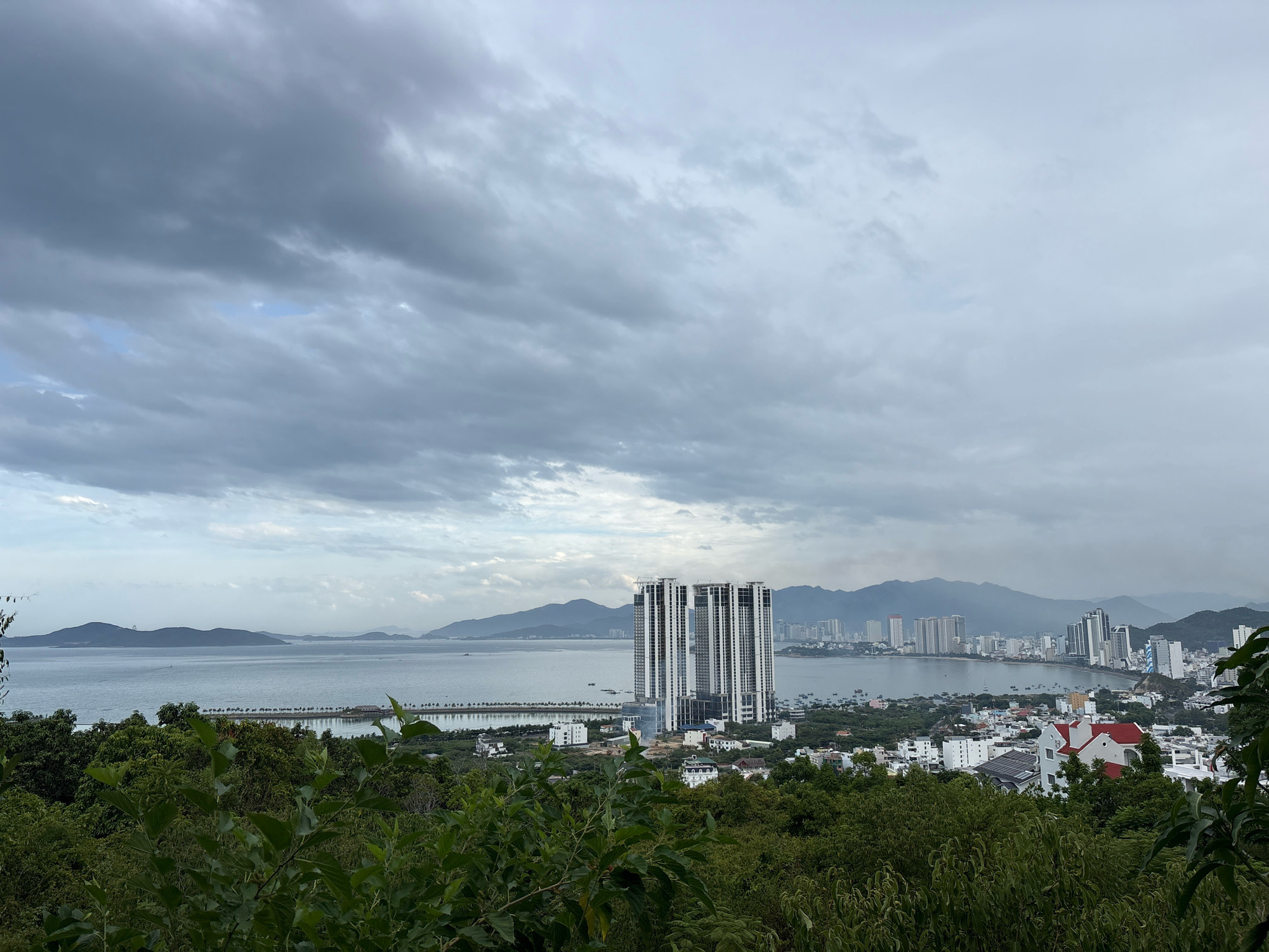 This is a wide-angle, elevated, daytime, landscape photograph of a coastal city, likely Nha Trang, Vietnam, under a cloudy sky. In the foreground, lush green trees and foliage dominate, framing the scene. Beyond the trees, a body of water, possibly the sea or a bay, stretches towards the horizon. Across the water, a cityscape is visible, featuring numerous buildings of varying heights, including several modern high-rise towers, particularly a cluster of four prominent, interconnected towers that appear to be under construction or recently completed. Further inland, a range of hazy, mountainous terrain forms the backdrop. The water is calm and reflects the overcast sky, with small boats dotting the surface. The overall impression is of a developing urban area nestled between a natural coastline and mountains.