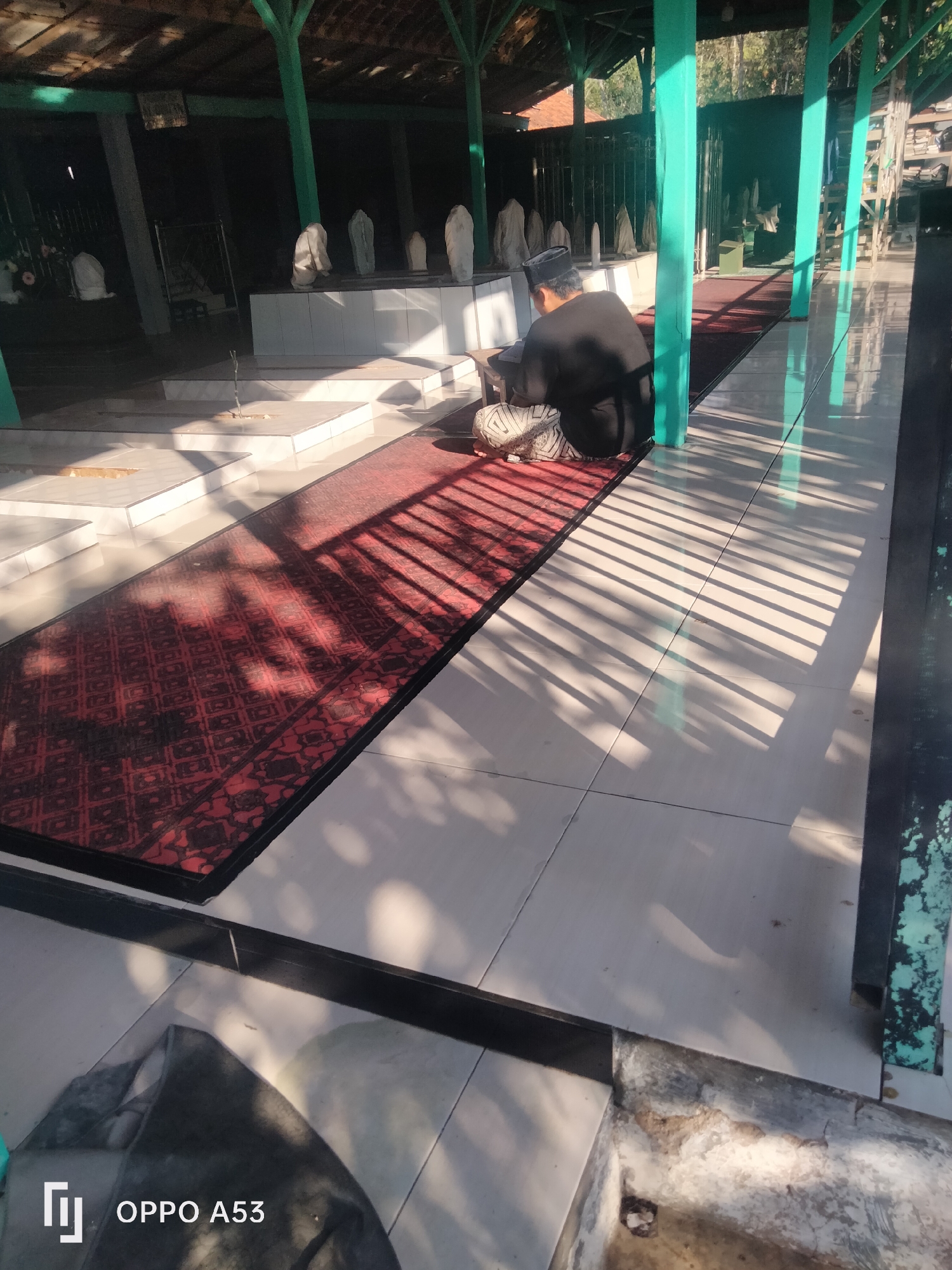 A man is sitting on a red rug, facing away from the camera, in a covered outdoor area. He is wearing a black shirt and a black peci hat, and has a striped cloth wrapped around his waist. He is looking down at something in front of him. In the background, there are several white grave markers. The ground is tiled, and there is a red rug with a diamond pattern running down the middle of the tiled area. Sunlight is casting strong shadows through the poles of the shelter. The scene appears to be a cemetery or a burial site.