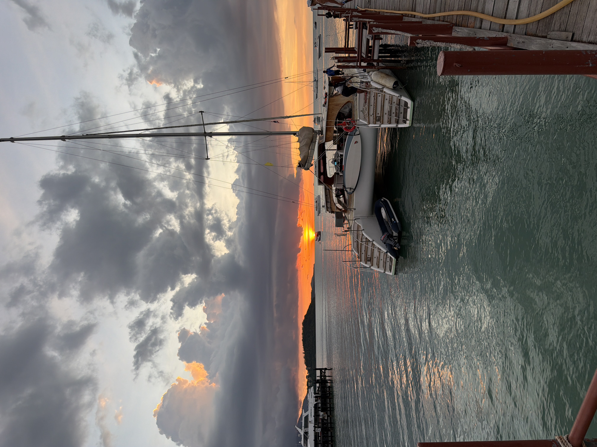The image depicts a sunset scene at a pier in Baan Bang Rak, Thailand. A large catamaran sailboat is docked at the pier, with its mast prominently visible against the dramatic sky. The sky is filled with dark, heavy clouds, but breaks of orange and yellow light from the setting sun illuminate the scene, casting reflections on the calm, greenish water. Several other boats, including a larger white yacht, are visible in the distance, anchored or docked. Two people are standing on the pier near the catamaran, and another person is visible on the far right of the pier. The pier itself is made of weathered wooden planks, with red posts and mooring ropes. The overall atmosphere is serene and picturesque, capturing the natural beauty of the location at dusk.