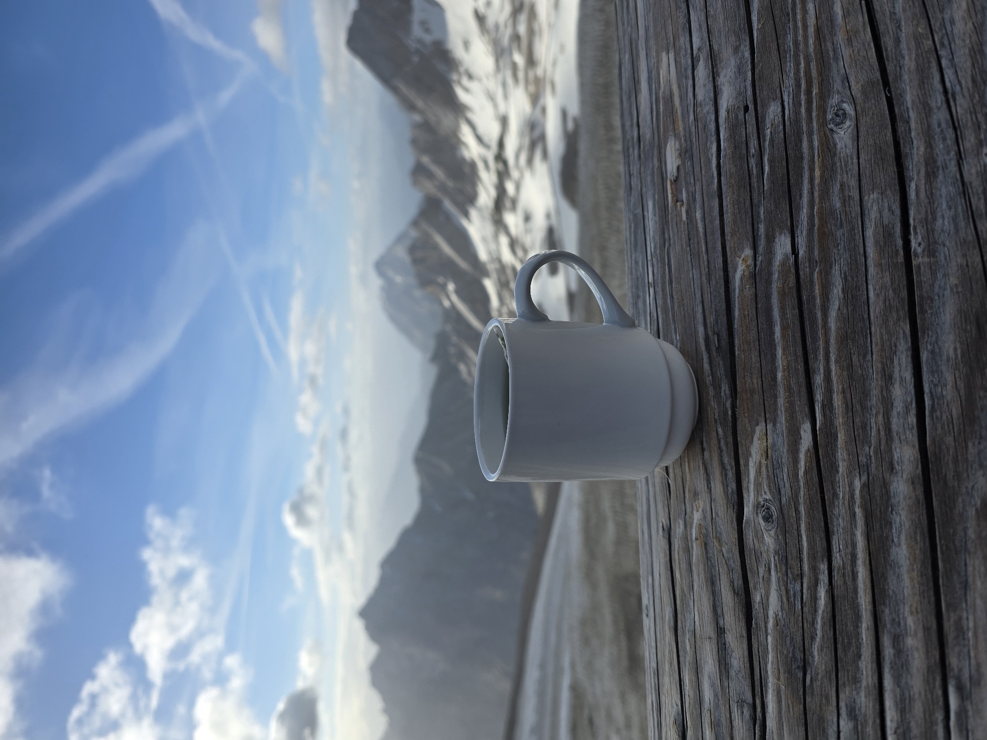 A white mug with a dark liquid sits on a weathered wooden surface. The background features a panoramic view of snow-capped mountains under a blue sky with wispy clouds. The scene appears to be set in a high-altitude location, possibly near the Zugspitze, Germany's highest peak, which is in the vicinity of Garmisch-Partenkirchen. The lighting suggests it is daytime, likely morning or afternoon. There are no people visible in the image.