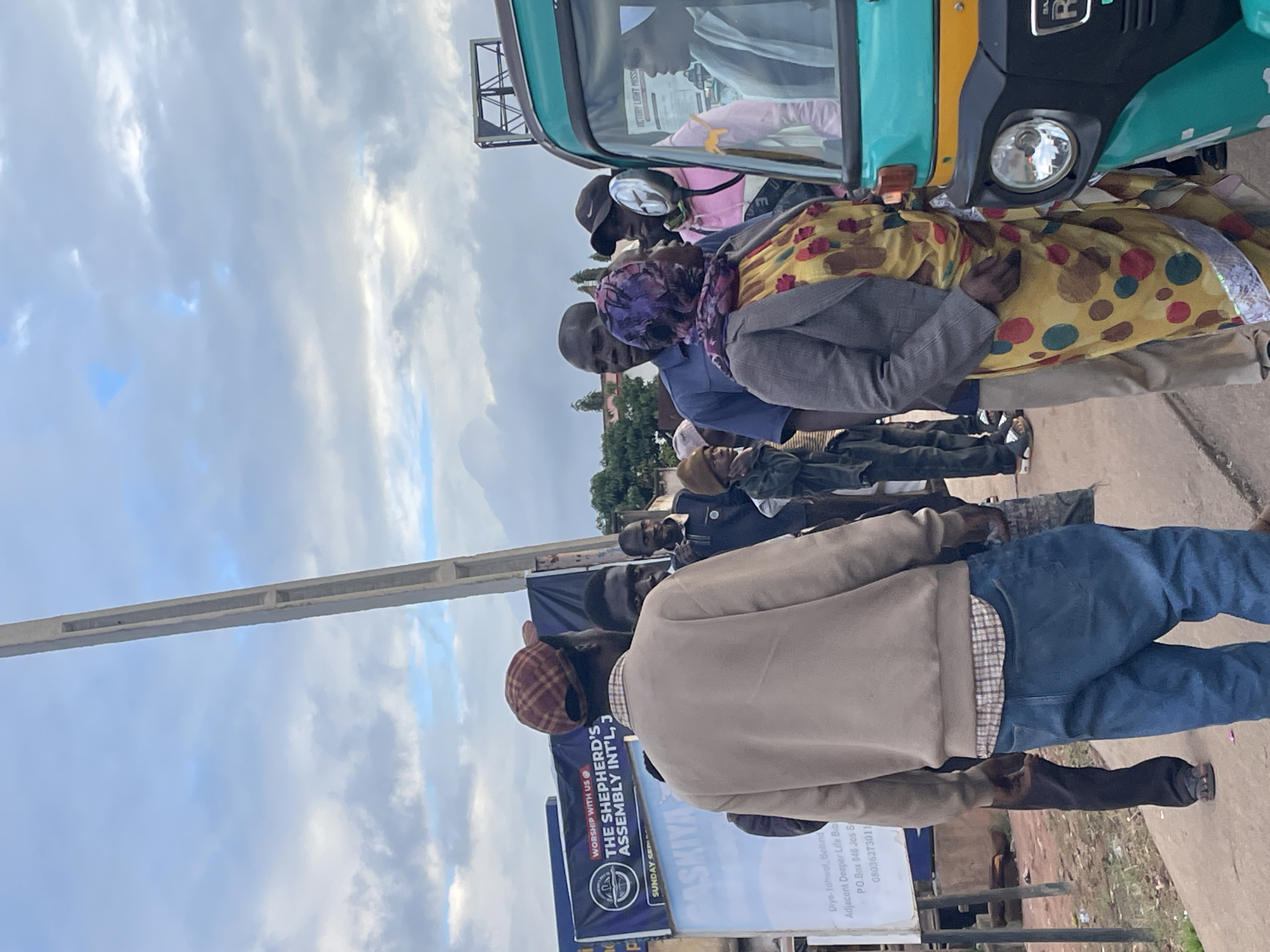 The image depicts a street scene in Jos, Nigeria, with a group of people gathered near a teal-colored auto-rickshaw. A tall, white concrete pole is visible on the right side of the frame, extending towards the cloudy sky. Several individuals are standing around, some appearing to be in conversation.
In the foreground, a person wearing a tan sweatshirt, a plaid collared shirt underneath, and blue jeans is facing away from the camera, with their back to the viewer. To their left, another person in dark pants is also visible.
To the right of the center, a woman in a gray jacket over a yellow, polka-dotted dress and a colorful headscarf is talking to someone inside the auto-rickshaw. A man in a dark blue shirt is standing behind her. Further to the right, a person in a pink hooded jacket is partially obscured. Inside the auto-rickshaw, a woman wearing a white veil is seated.
In the background, a large blue banner with white text is displayed, including "WORSHIP WITH US @ THE SHEPHERD'S ASSEMBLY INT'L, J..." and "SUNDAY SERV...". Another white sign with text, including an address and phone number, is visible to the left of the banner. The environment appears to be an urban setting with a paved walkway or road. The overall atmosphere seems to be a moment of informal gathering or interaction on a street.