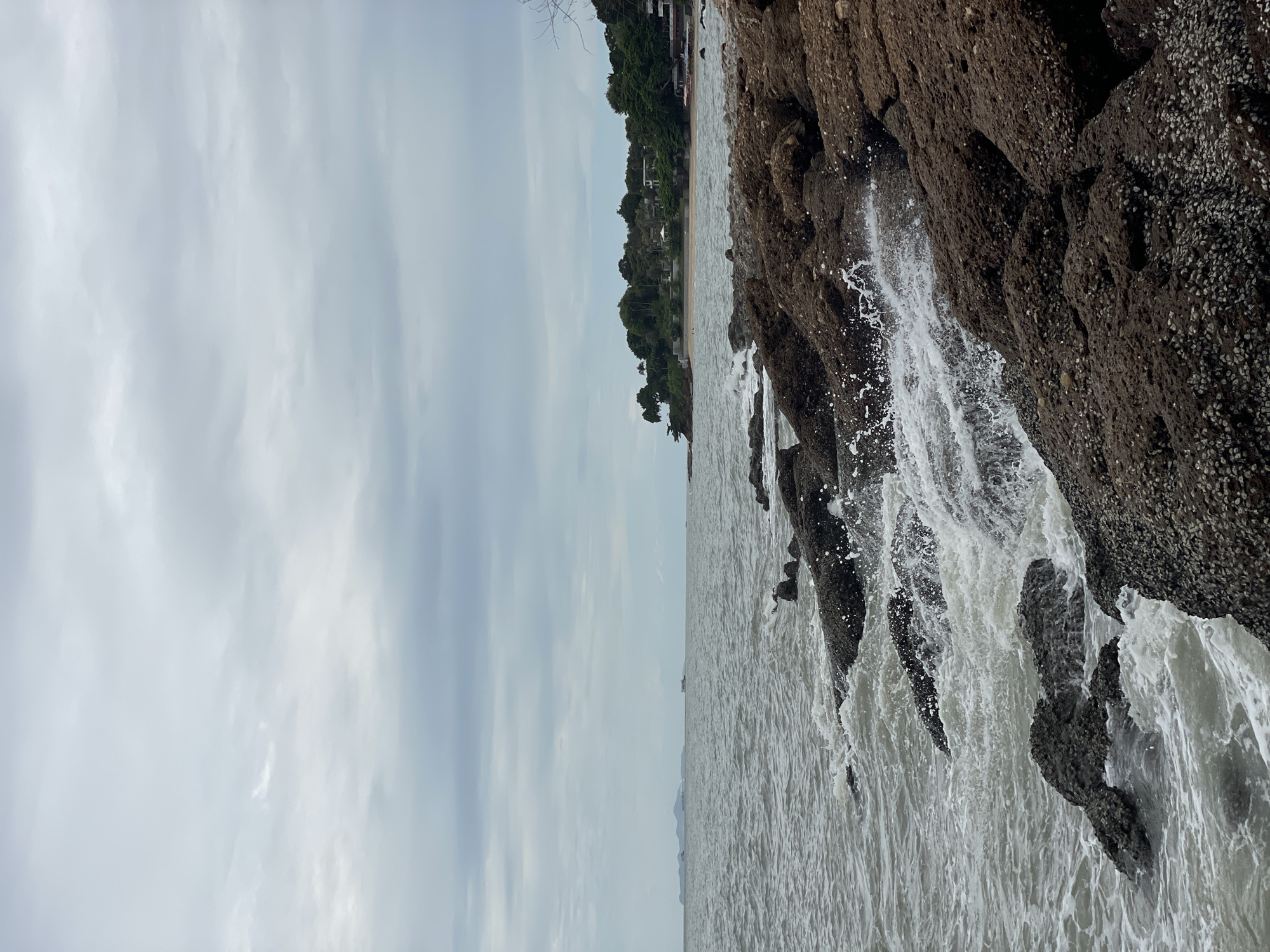The image depicts a rocky shoreline with the ocean waves crashing against the rocks. The sky is overcast with clouds. In the distance, there is a coastline with trees and some buildings, suggesting a developed area. The water is a muted gray-blue, and the rocks are predominantly dark brown and covered with barnacles or similar marine growth. The scene appears to be a natural coastal landscape within a tropical or subtropical region, likely in the vicinity of Pattaya City, Thailand, known for its beaches and coastal scenery. There are no people visible in the image.