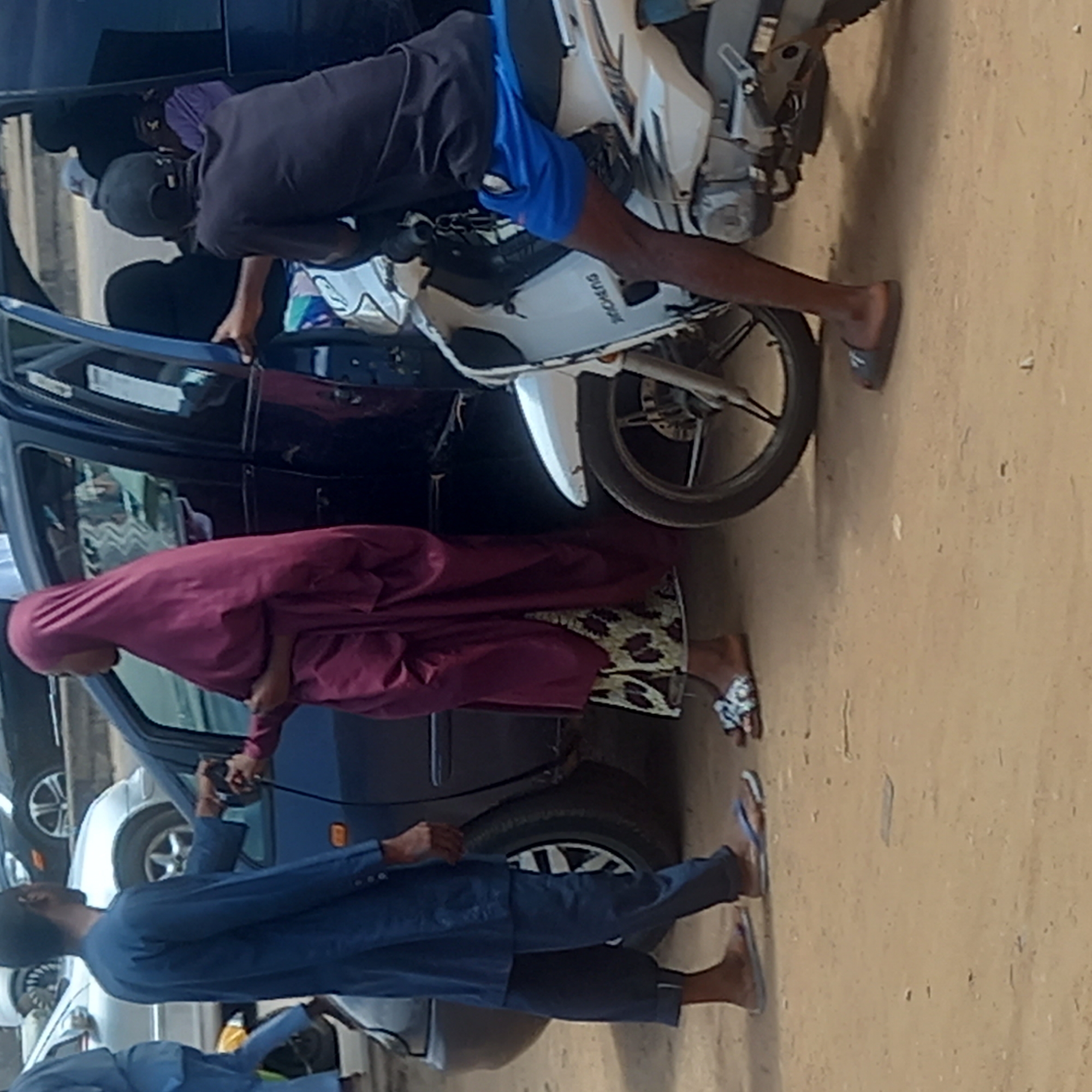 In Sardauna, Nigeria, a man in blue shorts and a black shirt sits astride a white motorcycle next to a dark blue car, leaning inside through the driver's side window. A woman in a maroon robe stands by the open passenger side door of the same car. To her left, a man in a dark blue suit holds a black object connected by a wire to the car. Other cars are visible behind the blue car. The ground appears to be a sandy or dirt surface.
