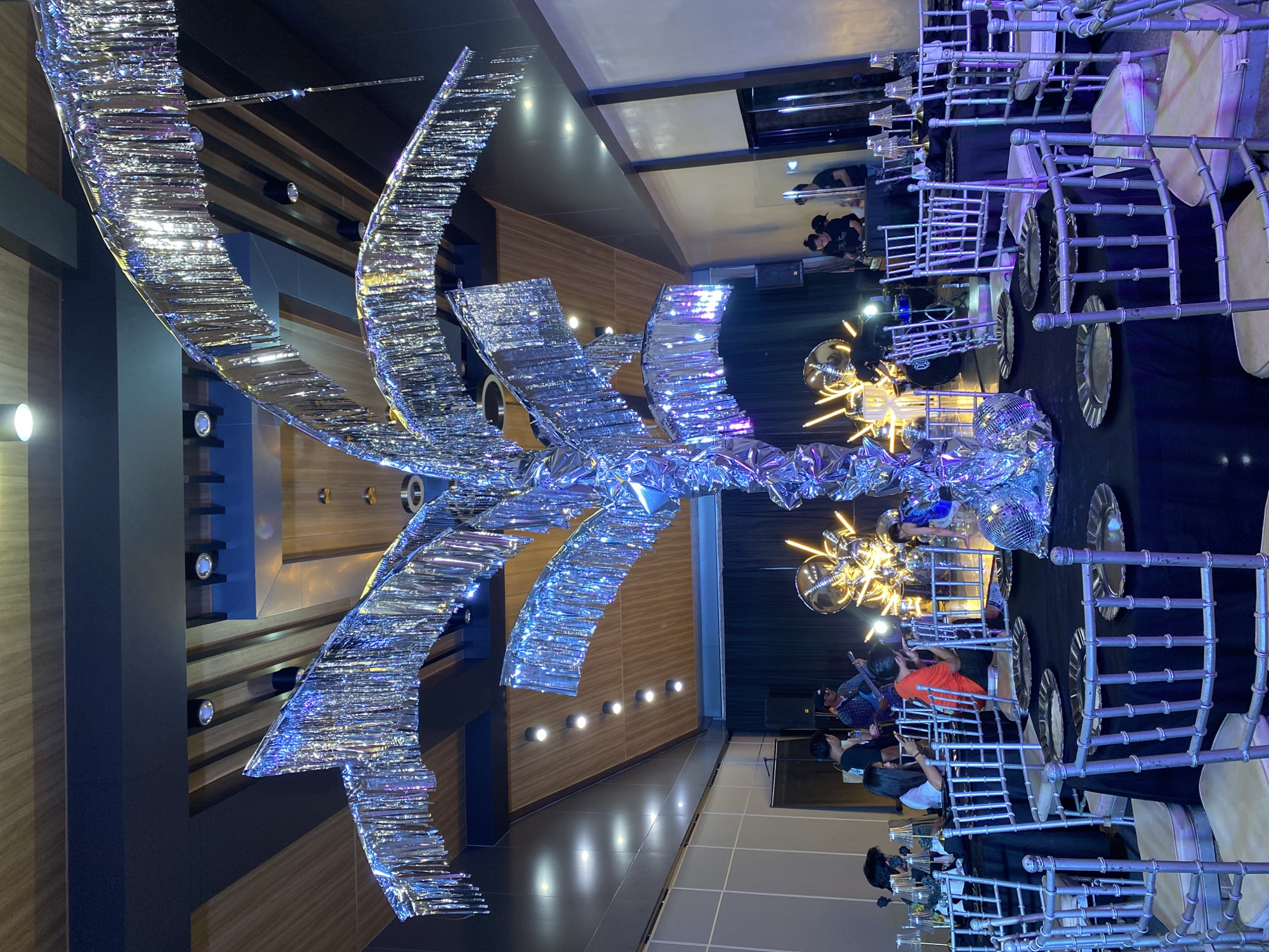 A group of people are gathered at tables set for an event, with a band playing music in the background. The room is decorated with a large, shimmering, palm tree-like centerpiece made of silver foil, and other metallic decorations. The overall atmosphere appears to be festive and celebratory. This scene is taking place at the The Royal Mandaya Hotel in Davao City, Philippines.