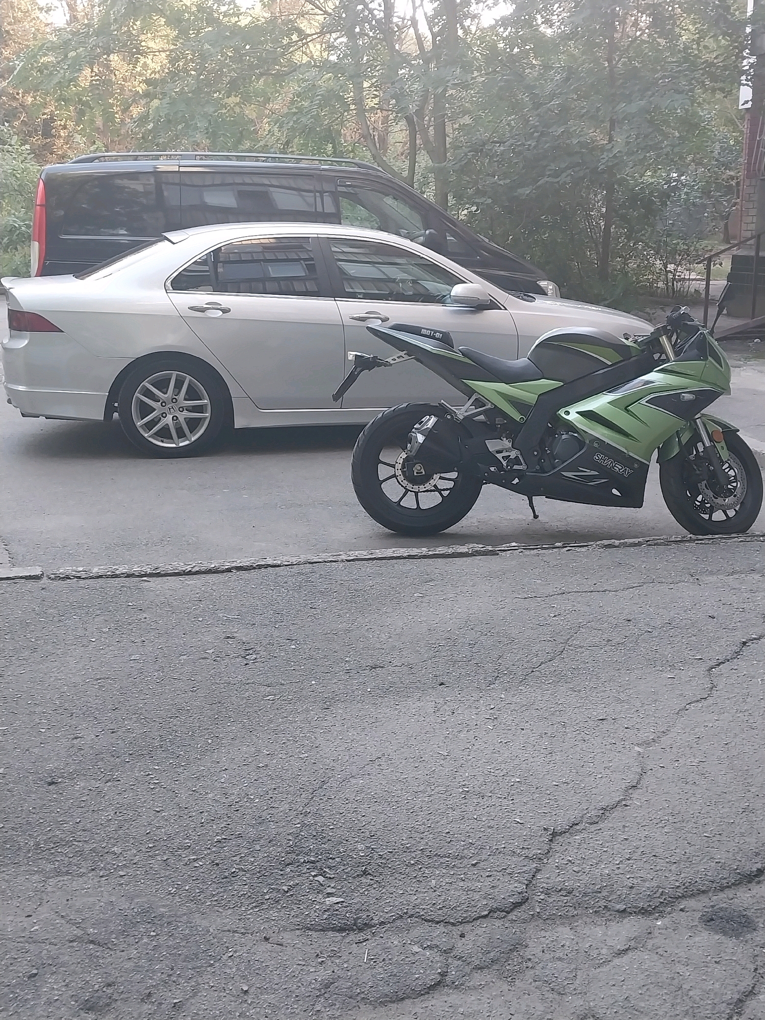 The image depicts a side-by-side view of a silver sedan and a green sport motorcycle parked on an asphalt surface. The sedan, partially visible, is parked behind the motorcycle. The motorcycle is the primary subject, positioned in the foreground, with its front tire slightly deflated. In the background, a black van is parked behind the sedan, and beyond that, dense foliage creates a natural backdrop. The scene appears to be a street or parking area within a city environment, likely Dnipro, Ukraine, given the context. The lighting suggests it is daytime, with soft light filtering through the trees. No people are present in the image.