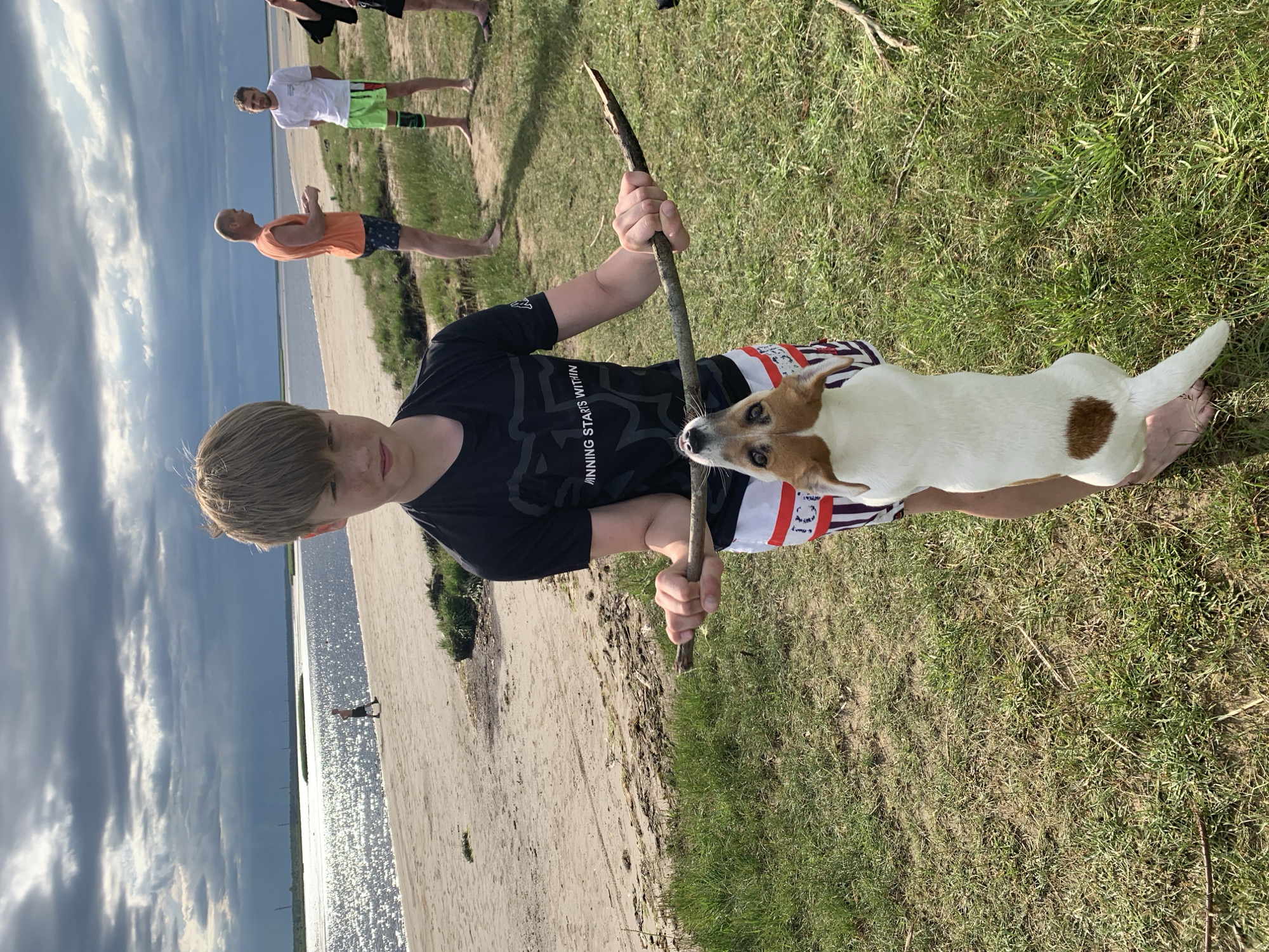 A boy is holding a stick horizontally across his body, with a small white and brown dog on its hind legs, holding onto the stick with its mouth. The boy is looking directly at the camera with a neutral expression, while the dog is looking up at the stick. The scene takes place on a grassy area near a body of water, likely a beach or riverbank. In the background, there are several other people standing and walking, and the sky is cloudy. The overall lighting suggests it is daytime, possibly overcast.