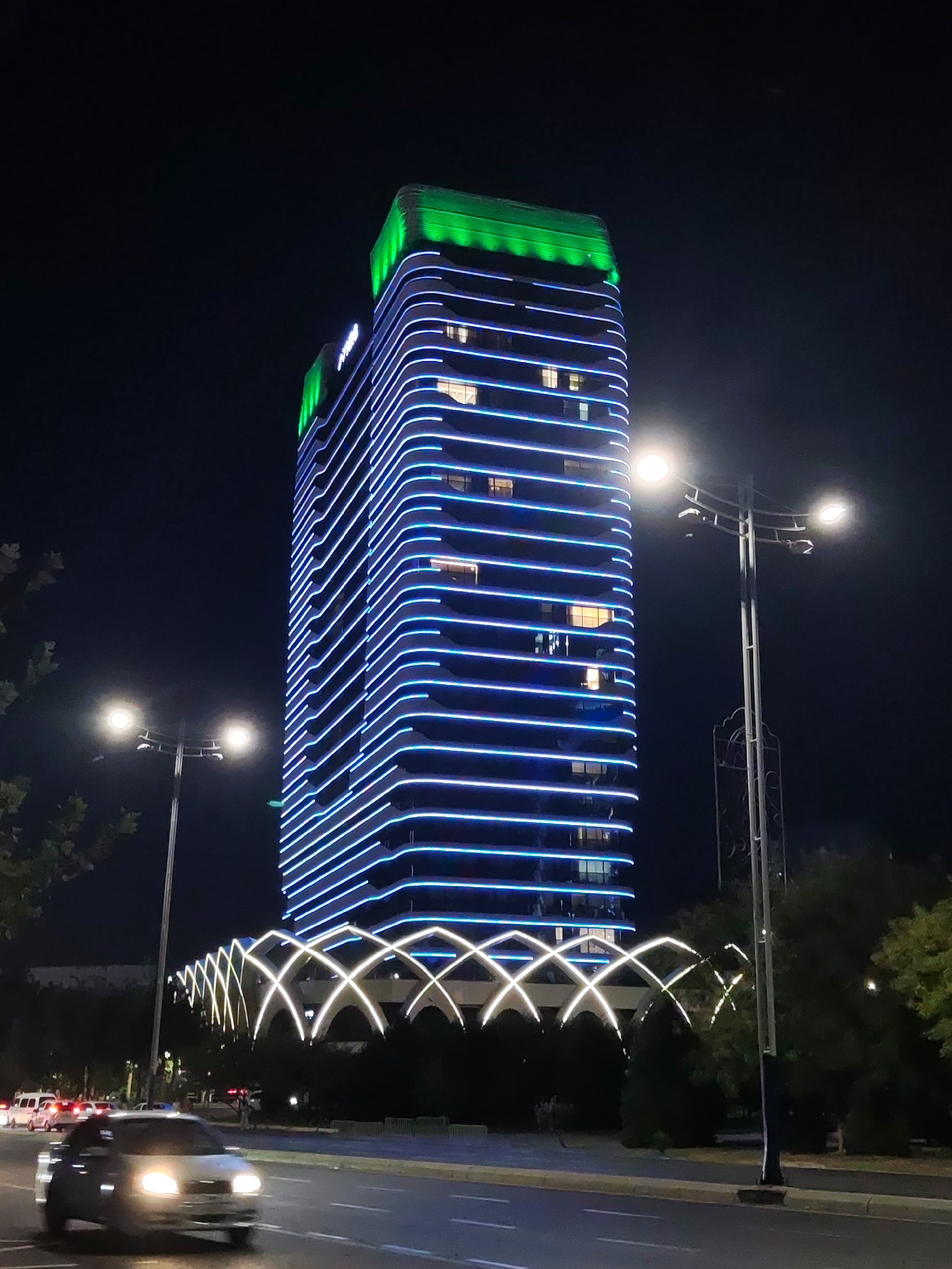 A modern, illuminated skyscraper stands tall against a dark night sky in Tashkent, Uzbekistan. The building is adorned with horizontal blue LED lights that create a striped pattern along its facade, with a green illuminated top section. In the foreground, a multi-lane road is visible with vehicles, including a silver car in motion and several other cars further down the road. Streetlights illuminate the area. In front of the skyscraper, a decorative archway with white and yellow lights creates an elegant entrance. Trees and foliage surround the base of the building and line the roadside, contributing to the urban landscape. The scene captures the vibrant nighttime ambiance of the city.