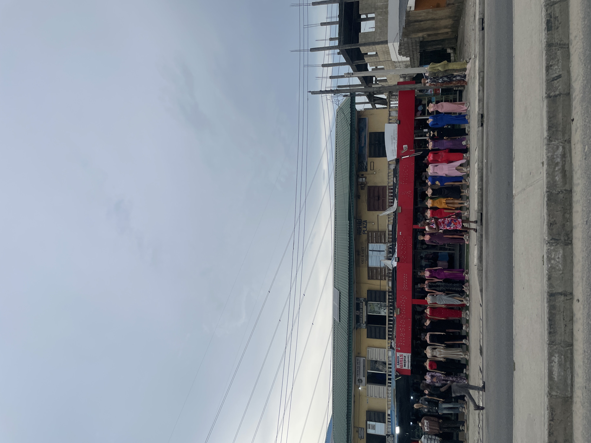 The image captures a street scene in Ado, Nigeria, depicting a commercial area with shops and buildings, some under construction. The photo is taken from the street level, looking towards a row of establishments.
**Subjects:** The foreground features a sidewalk next to a road. Along the sidewalk, numerous mannequins are displayed, showcasing a variety of colorful dresses and outfits. A few individuals can be seen walking on the sidewalk, with one person in the lower right walking past the mannequins. In the background, there are multi-story buildings. One building, possibly a plaza, has a yellow facade with a green roof, and a prominent red storefront below. Adjacent to it, an unfinished concrete structure with exposed rebar indicates ongoing construction. Power lines crisscross the upper portion of the sky.
**Activity or event taking place:** The primary activity depicted is retail display and pedestrian movement. Mannequins are arranged to attract shoppers, while people are walking along the street, likely engaged in daily commutes or shopping.
**Notable details:** The time of day appears to be daytime, possibly late afternoon or early evening, under an overcast or partly cloudy sky, as suggested by the soft, diffused light and the gray tones of the clouds. The architecture, including the unfinished concrete building and the style of the commercial structures, is characteristic of urban areas in developing regions. The street appears to be paved, and the sidewalk is concrete.
**Visible text:** Several signs are visible on the buildings. On the red storefront, part of a sign reads "SALES GIRLS NEEDED". On the yellow building, signs that are partially legible include "AMIDU PLAZA", "OJE RASULY", and "OFFICIAL REYE".