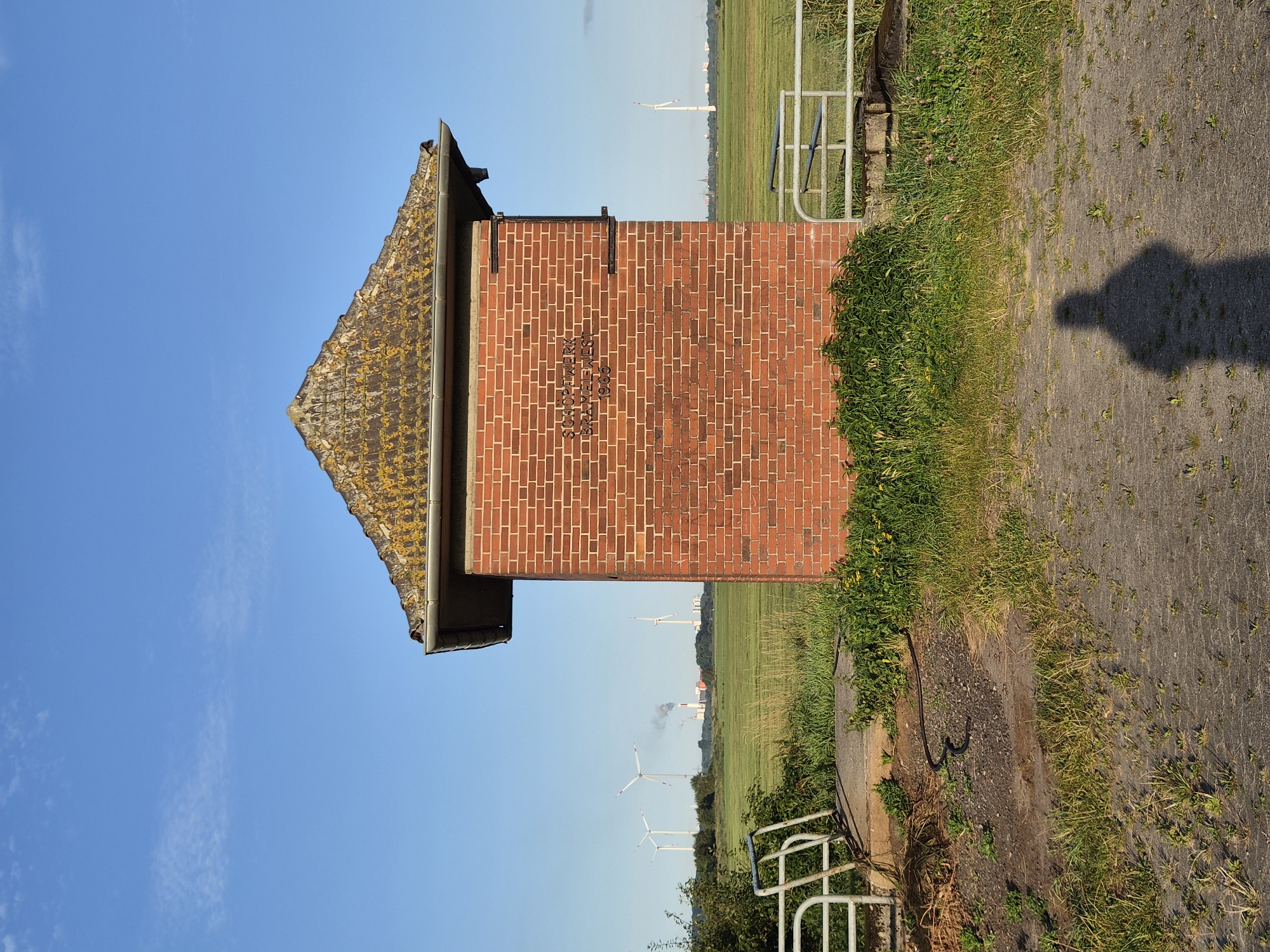 An exterior, eye-level, full-shot photograph captures a red brick building with a weathered, moss-covered, pyramid-shaped roof under a clear blue sky. The building is centrally located in the frame, with a patch of gravelly ground in the foreground and a grassy field behind it. A shadow of a person, presumed to be the photographer, is visible on the ground in the lower right corner.

The brick building has dark lettering etched into its facade, appearing to read "SCHOPFWERK BRAMELWEST 1965". Metal brackets are affixed to the sides of the building. To the left of the building, a concrete structure leads to a white metal railing, partially obscured by lush green vegetation. To the right, another section of white metal railing is visible, also bordered by greenery.

In the background, the grassy field stretches towards the horizon, dotted with several wind turbines. In the far distance, industrial structures with smokestacks emit plumes of smoke, and additional wind turbines are scattered across the landscape. The overall environment suggests a rural or semi-industrial setting. The scene appears to be located in Schiffdorf, Germany, as indicated by the building's inscription.