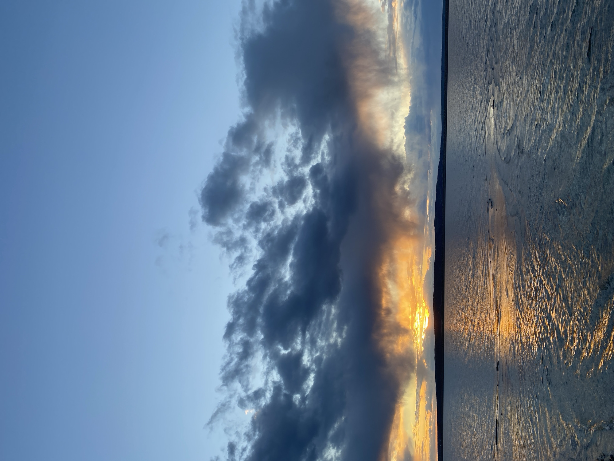 The image depicts a sunset over a body of water. The sky transitions from a clear blue to dark, ominous clouds highlighted by the golden light of the setting sun. The water reflects the sunlight, creating a shimmering effect. The location appears to be near a shoreline, possibly in Chaikovsky, Russia, given the prompt context, but there are no specific landmarks or features to definitively confirm this. No people or distinct objects are visible, focus is only given to the clouds and reflecting light on the water.